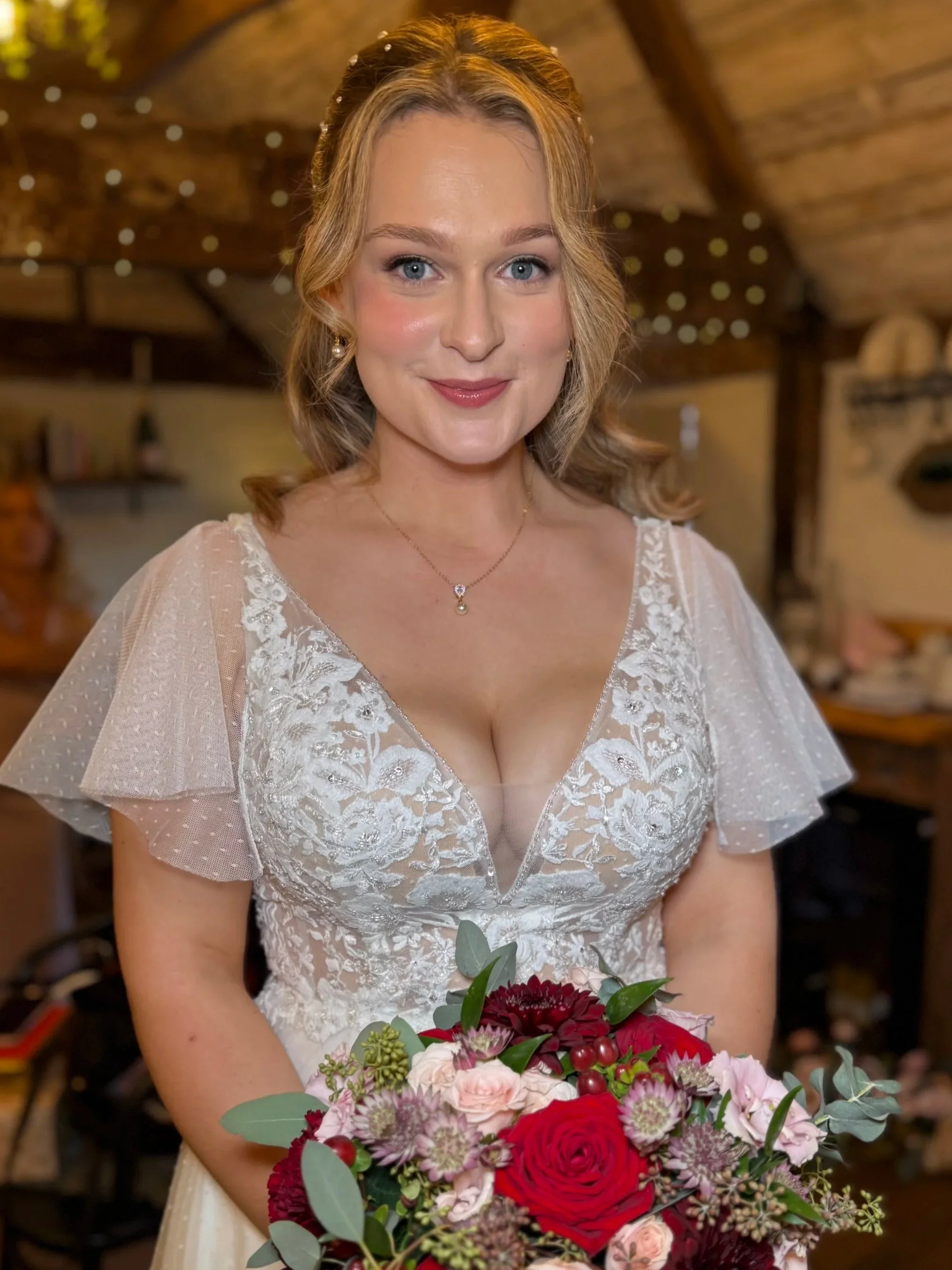 A woman in a wedding dress holding a bouquet of red, pink, and purple flowers, smiling in a rustic indoor setting.