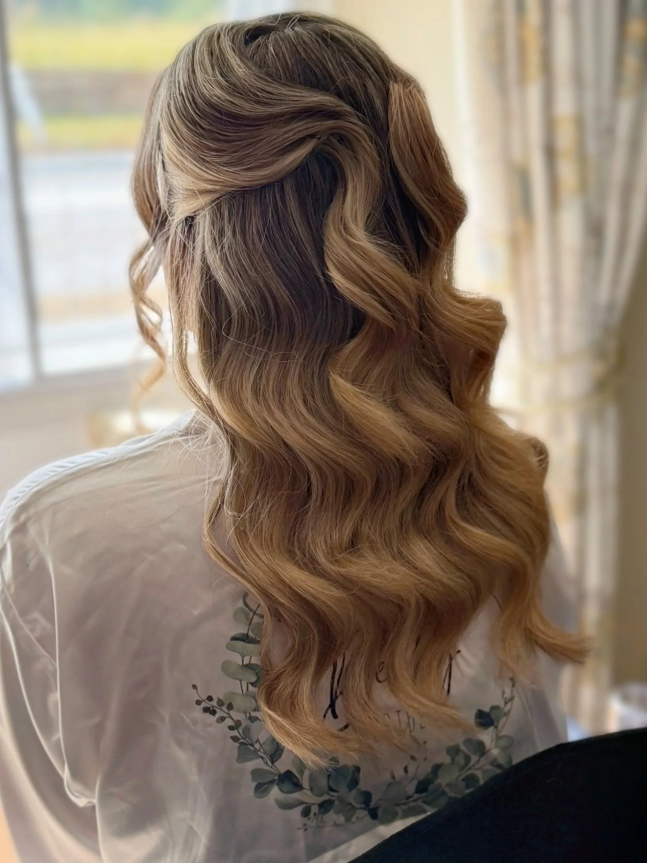 Back of a woman's head showing long, wavy hair with highlights, standing indoors near a window with blurred curtains.