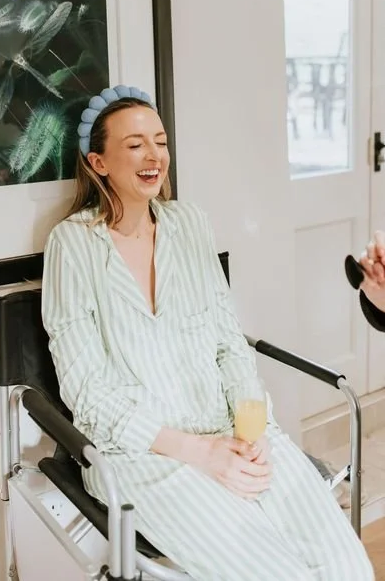 Young woman in hospital bed wearing pajamas and a blue headband, holding a glass of orange juice, smiling and talking to someone.