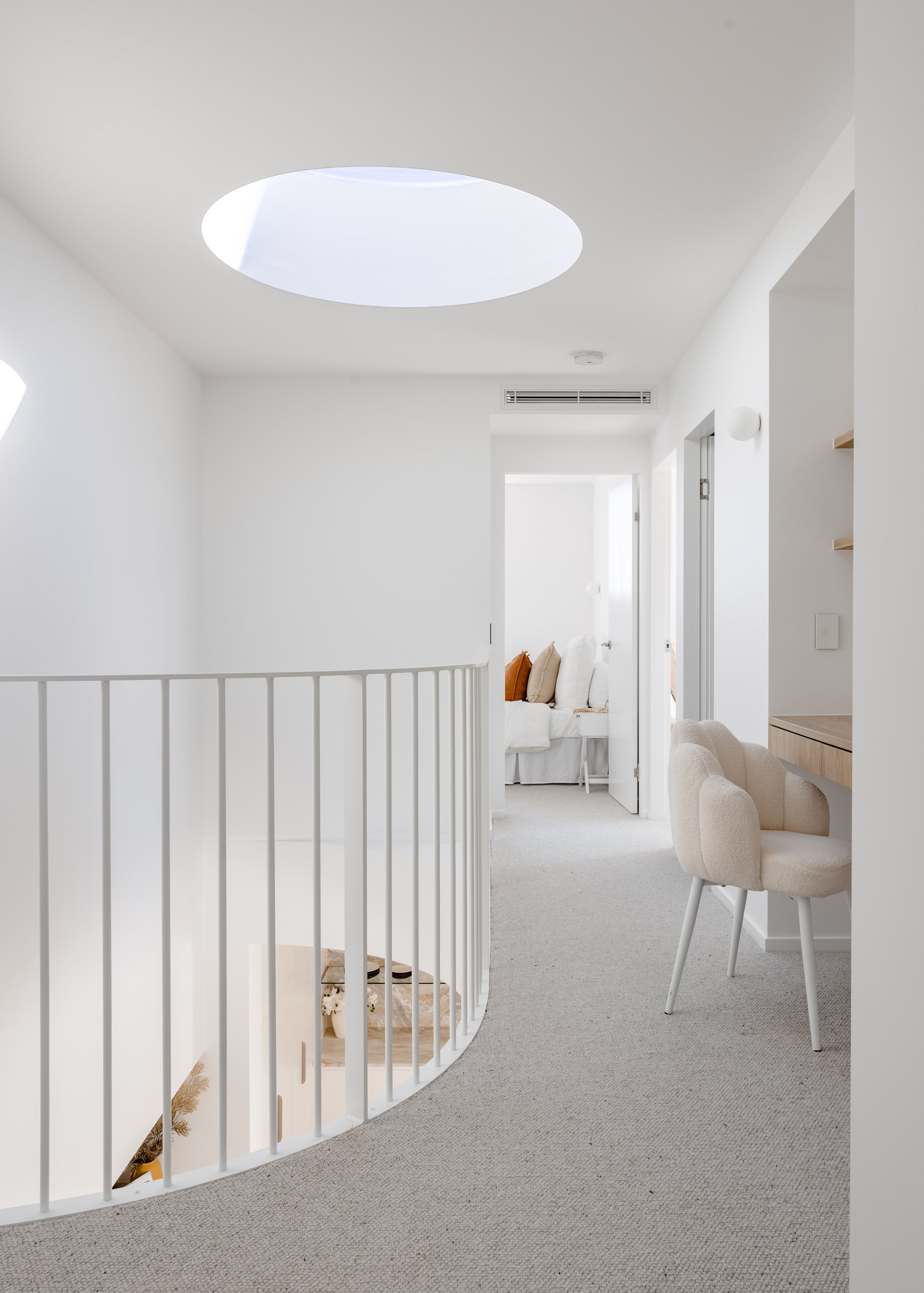 Bright, minimalist hallway with white walls and ceiling. Circular skylight and open doorways lead to a sitting area with white furniture and brown accent pillows. Beige carpet and modern chairs near a built-in desk or shelf.