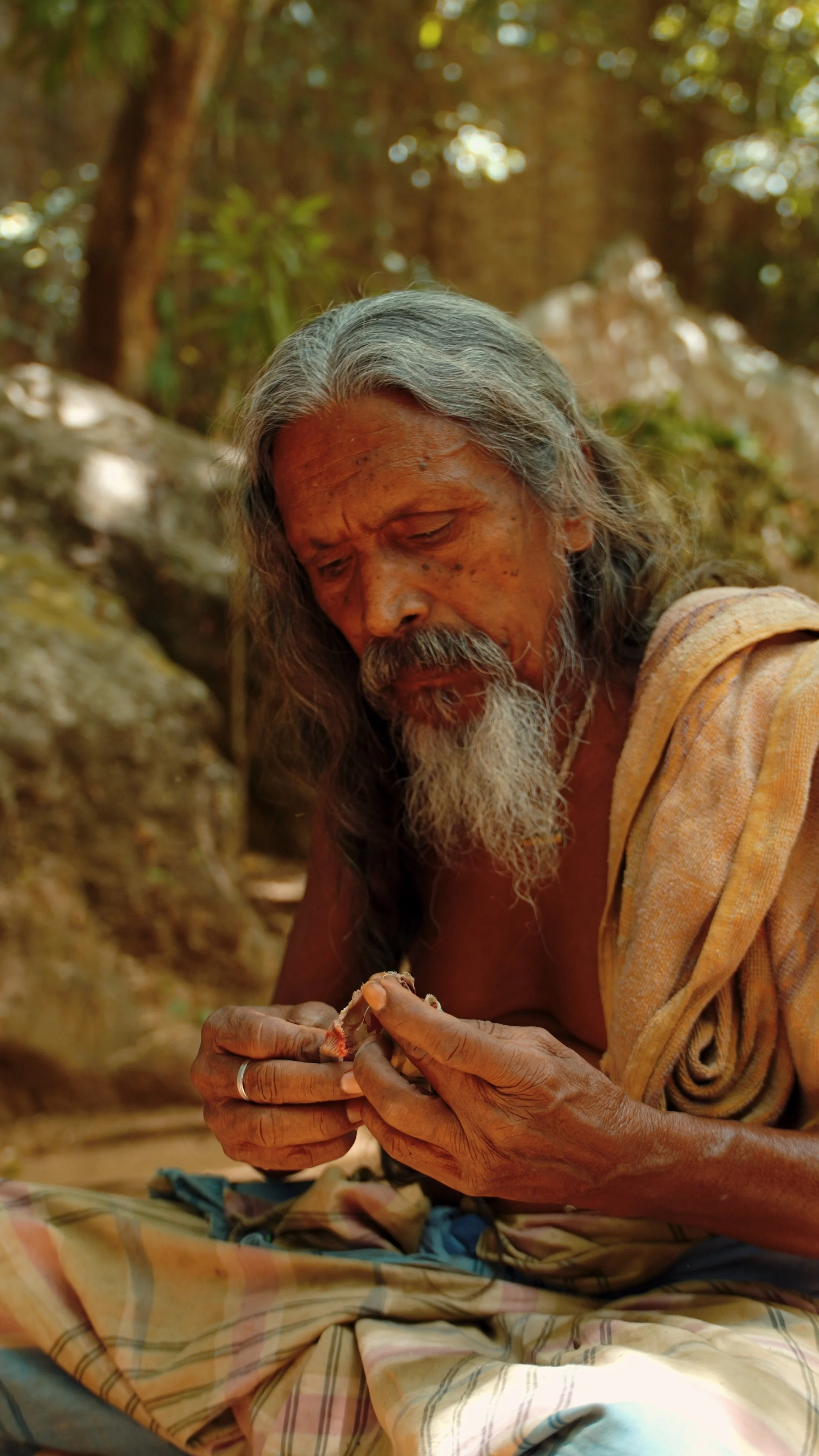 An elderly man with long gray hair and a beard, wearing a yellowish shirt, is sitting outdoors in a forested area. He is focused on an object in his hands, possibly a small animal or craft, with a thoughtful expression.