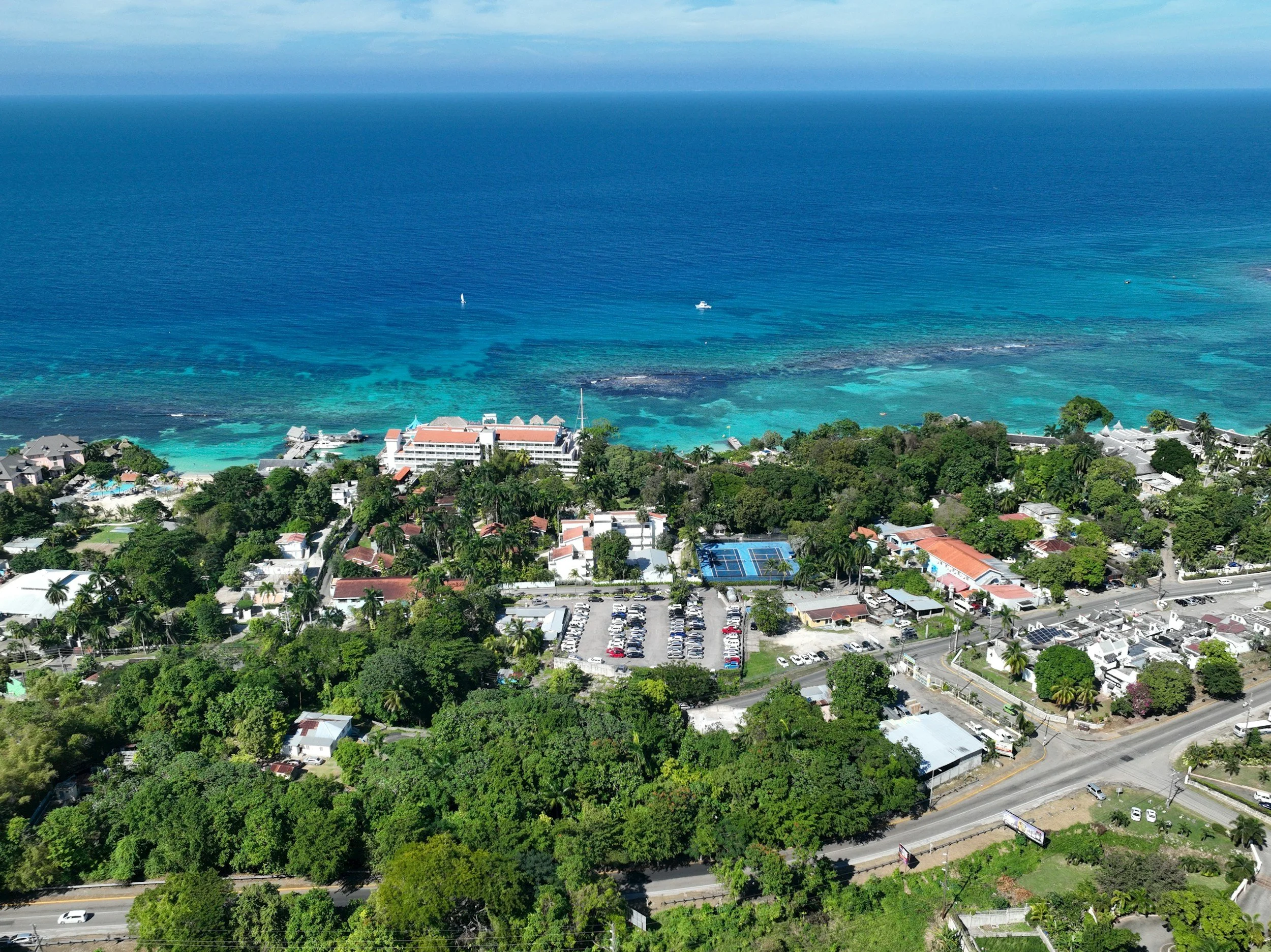 An aerial view of a coastal town with buildings, trees, and a parking lot near the shoreline, with clear blue ocean water and boats in the water.