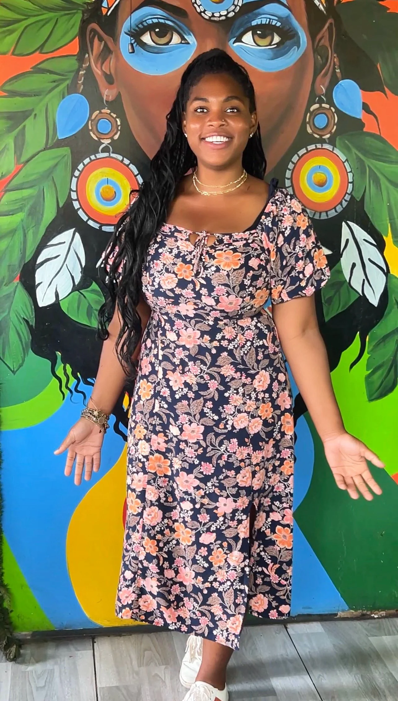 A woman with long, dark, curly hair and a cheerful expression poses in front of a vibrant mural of a woman's face with large eyes, surrounded by green leaves and colorful circles. She is wearing a floral dress, white sneakers, and jewelry.
