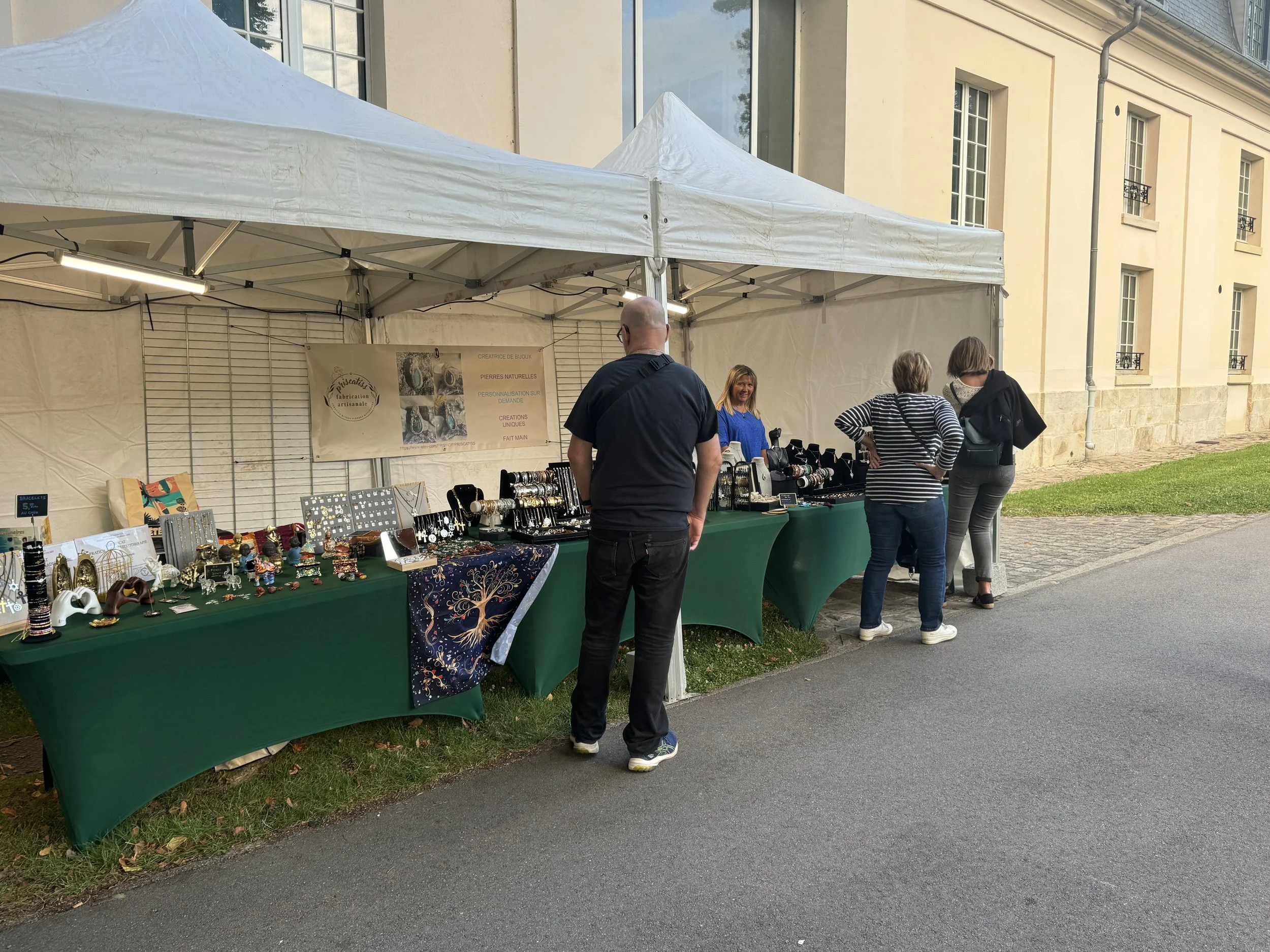 Stand artisanal avec bijoux et objets décoratifs, plusieurs personnes regardant les produits, sous une tente blanche à côté d'un bâtiment ancien.