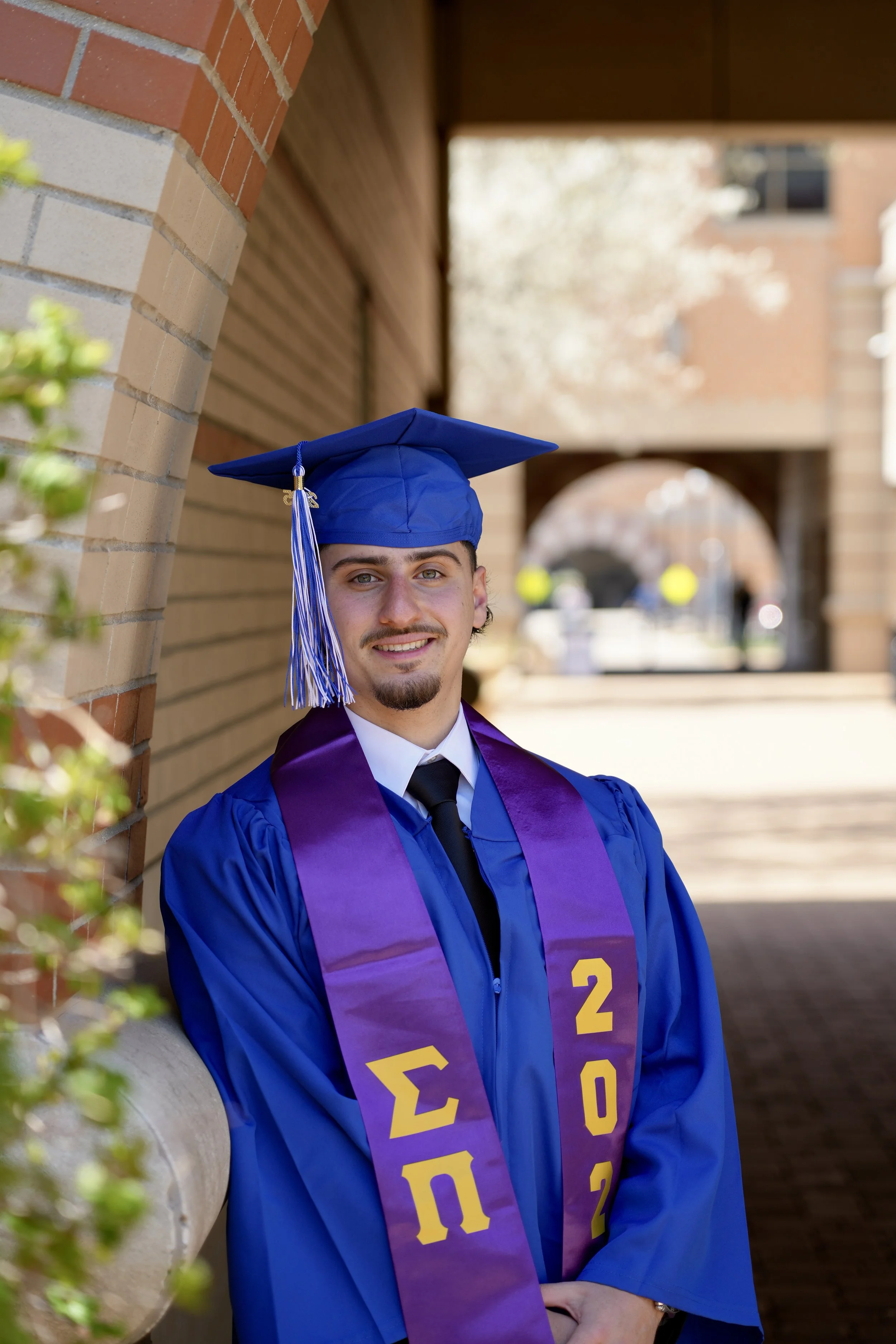 Young man in blue graduation gown and cap with purple stole standing outdoors near a brick wall, smiling at the camera.