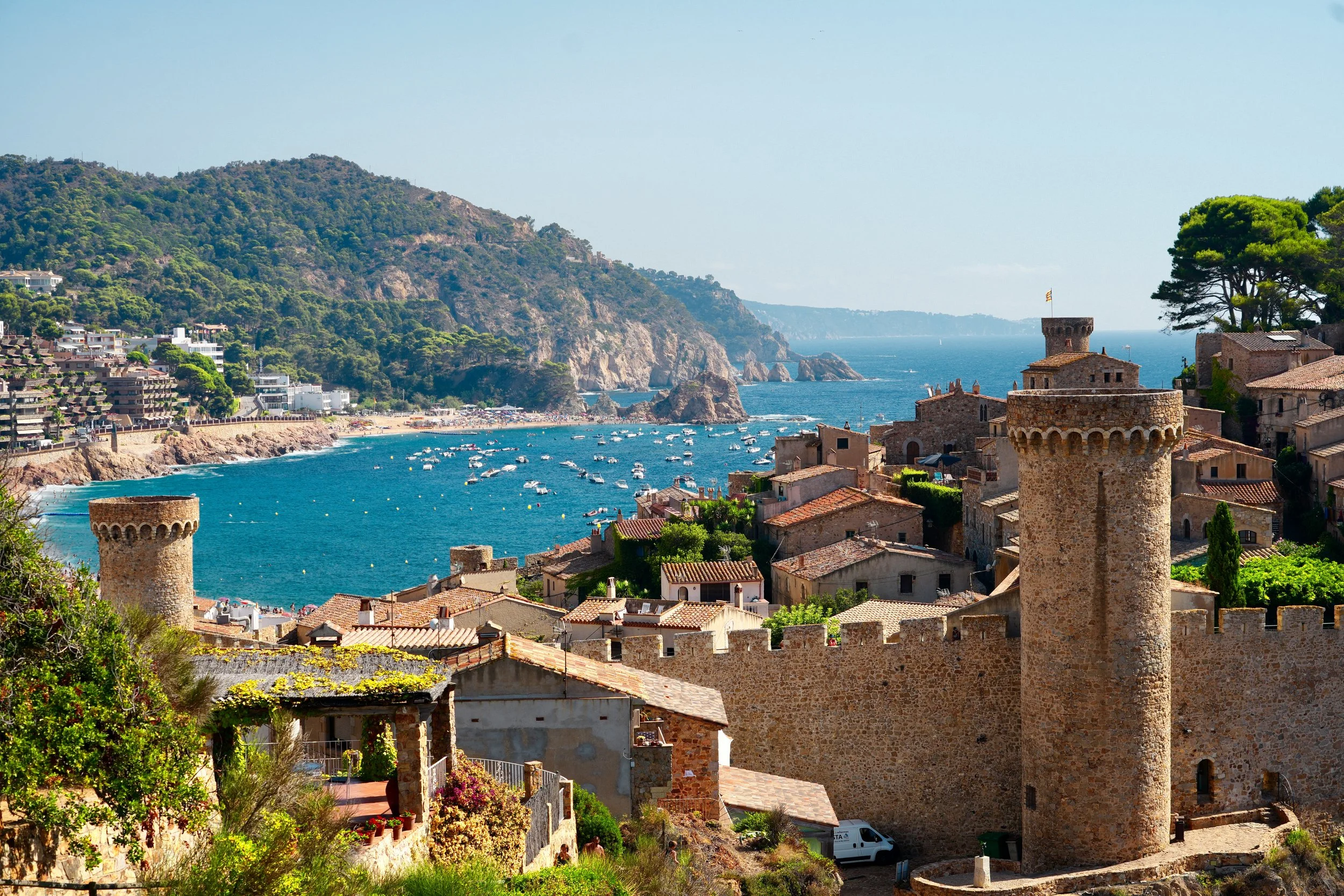 Medieval stone towers and walls overlooking a coastal town with blue sea and hills in the background.