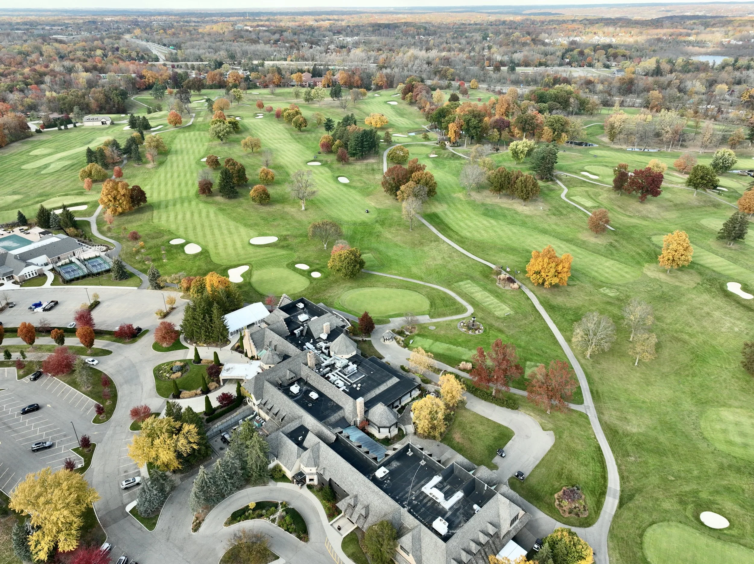 Aerial view of a golf course with clubhouse and parking lot, surrounded by trees with autumn foliage.