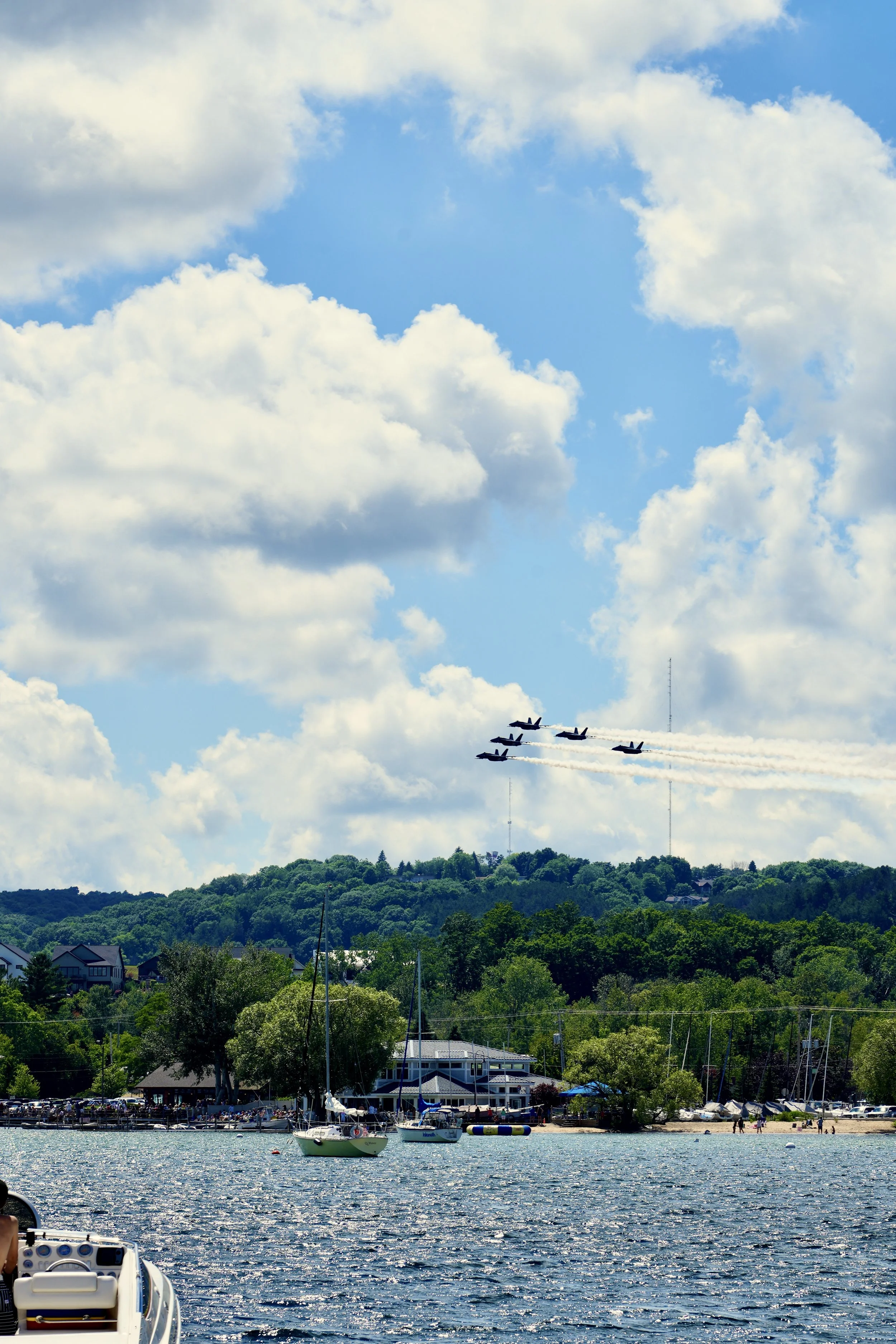 A formation of jets flying over a lakeside area with sailboats, trees, and buildings under a partly cloudy sky.