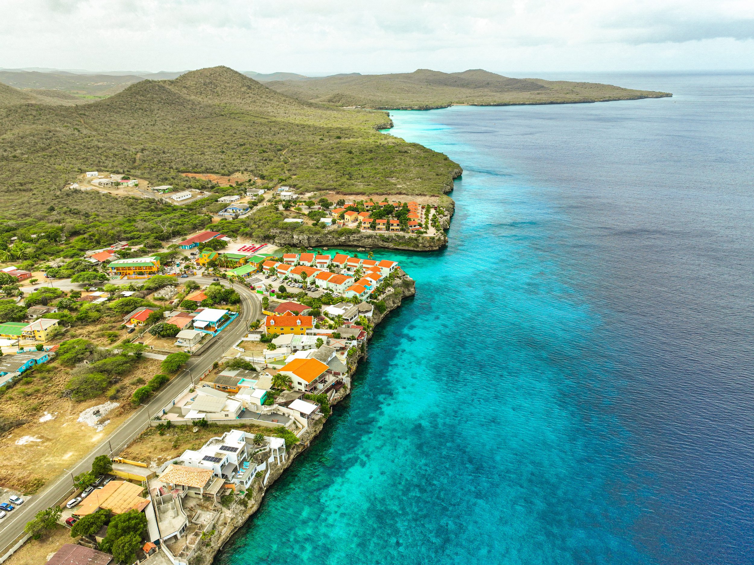 Aerial view of a coastal town with colorful buildings on a cliff overlooking turquoise ocean waters, surrounded by green hills and mountains in the background.