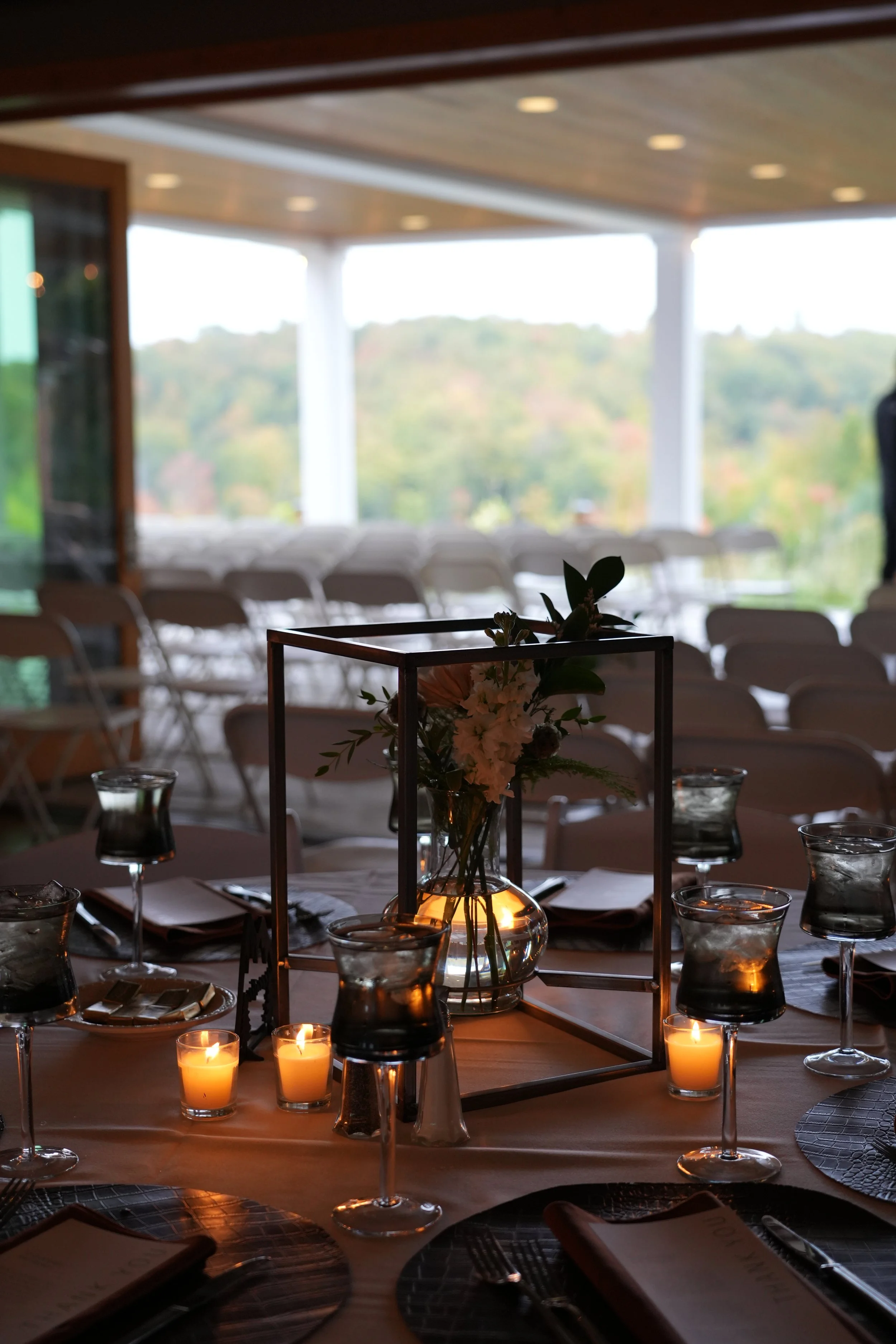 Elegant table setting with lit candles, wine glasses, and a floral centerpiece in a dimly lit venue with rows of chairs in the background.