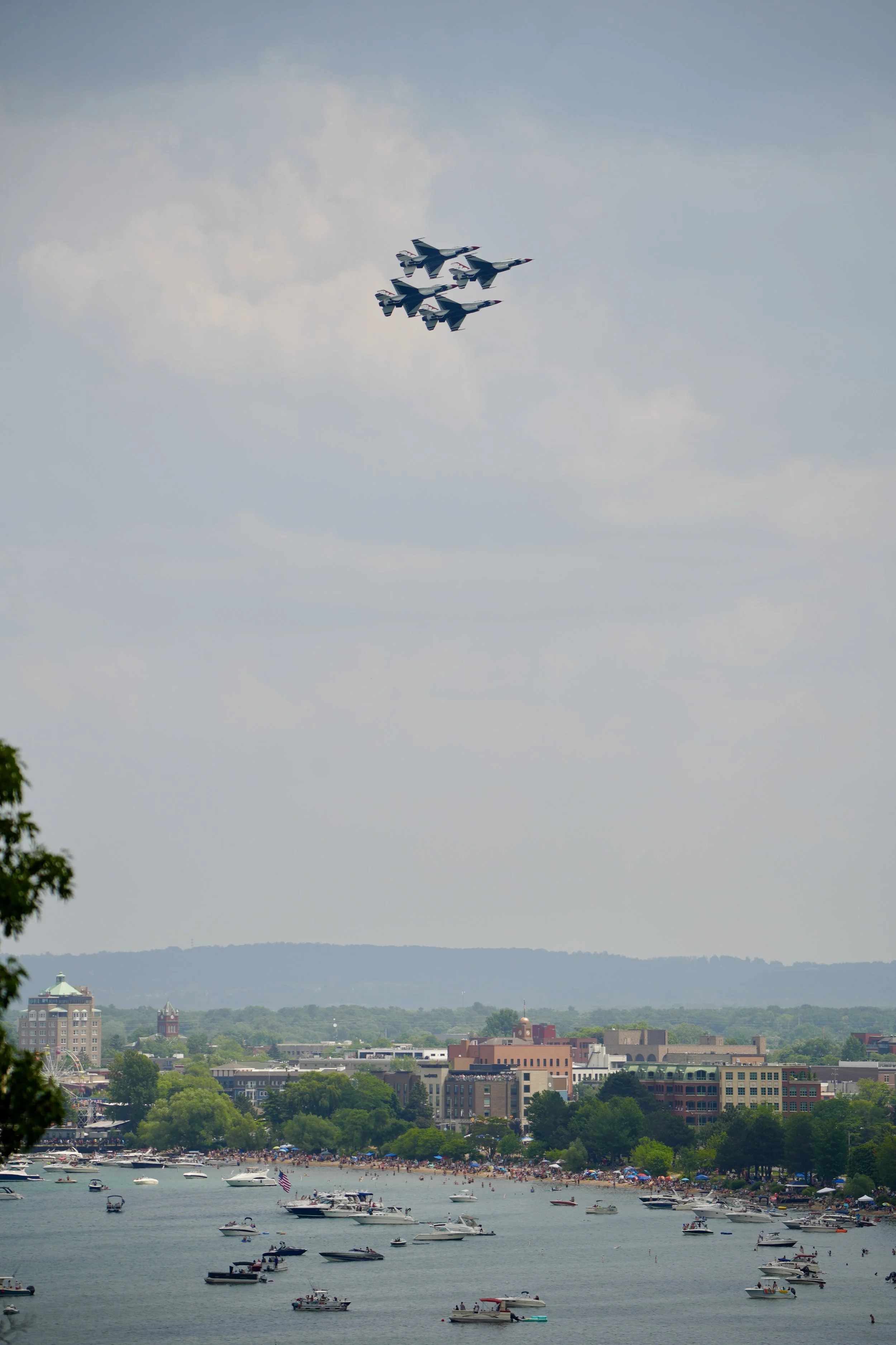 Four fighter jets flying in formation over a cityscape with numerous boats on the water below.