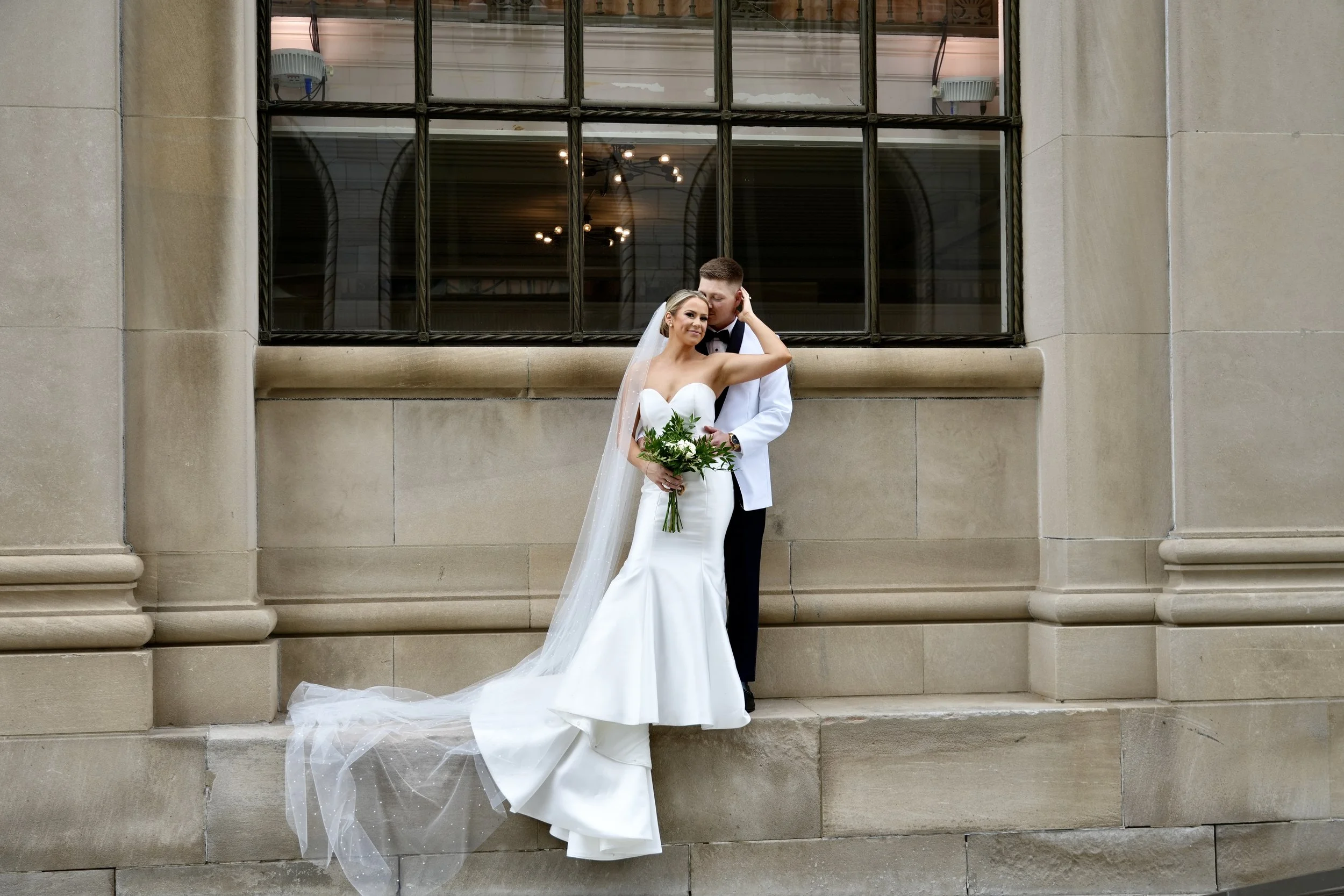 Bride and groom in formal attire pose against a building wall, with the bride in a white wedding dress holding a bouquet and the groom in a white tuxedo jacket.