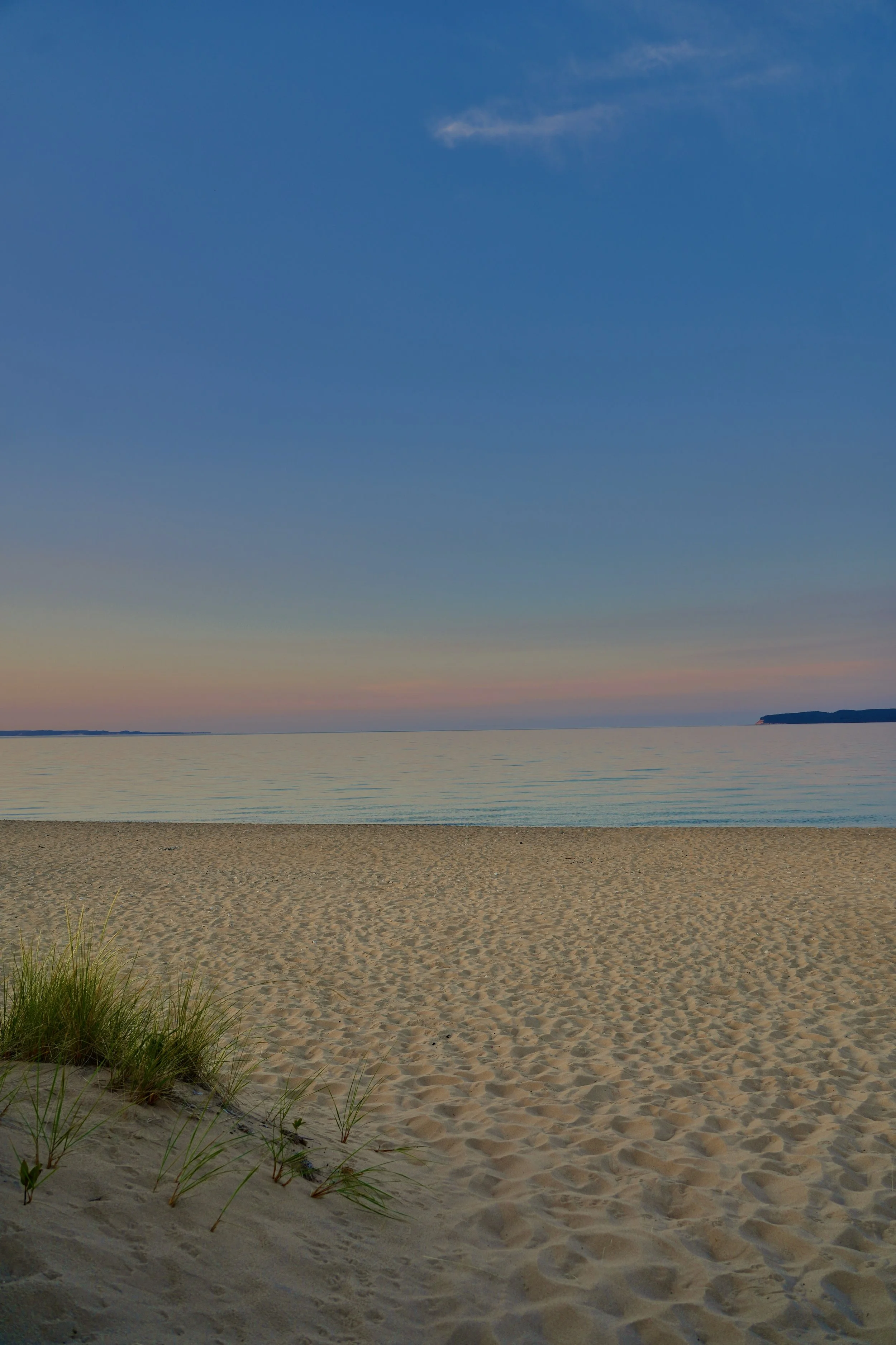 Sandy beach with grass tufts, calm ocean under a clear sky, horizon at sunset.