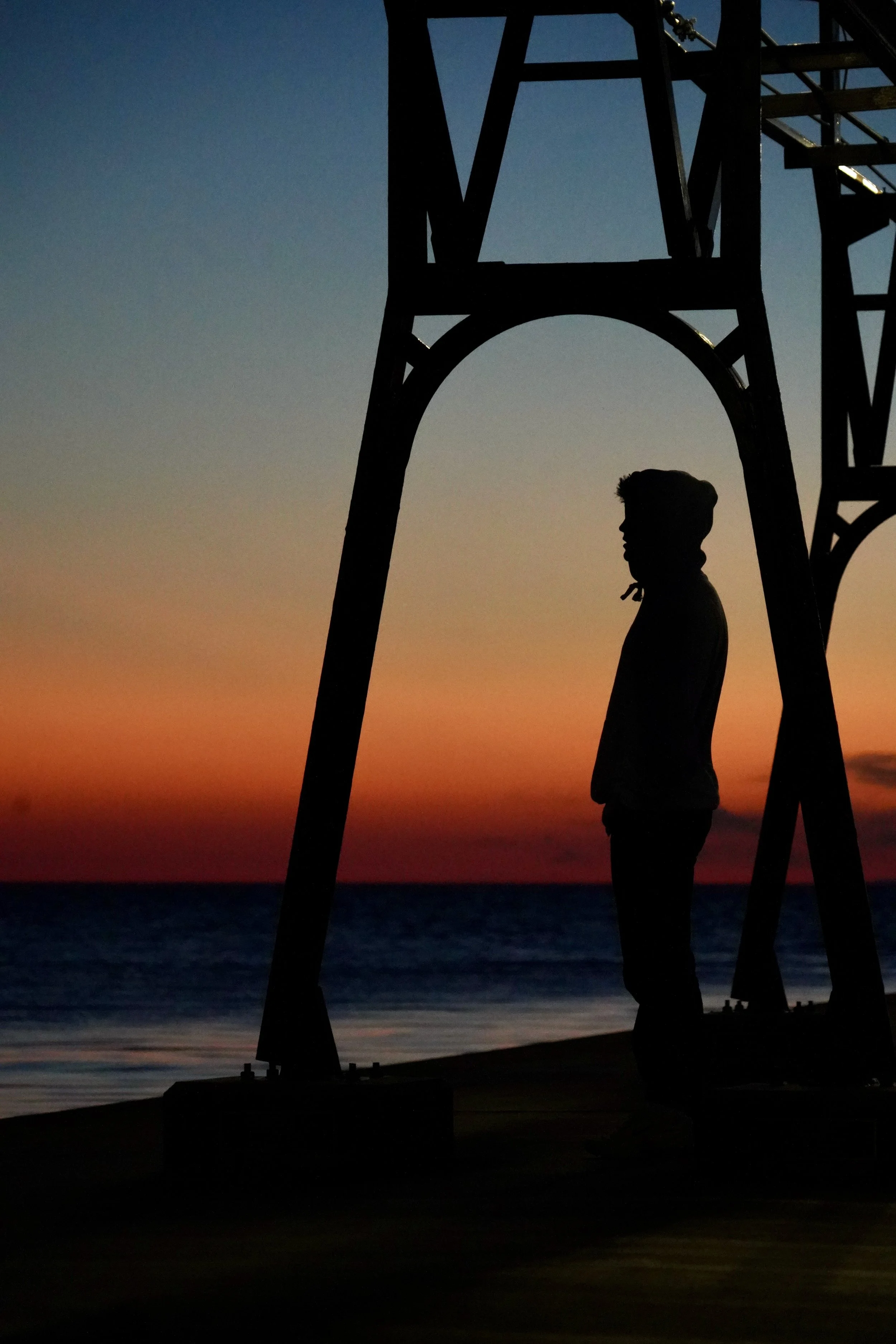 Silhouette of person under structure at sunset by the ocean
