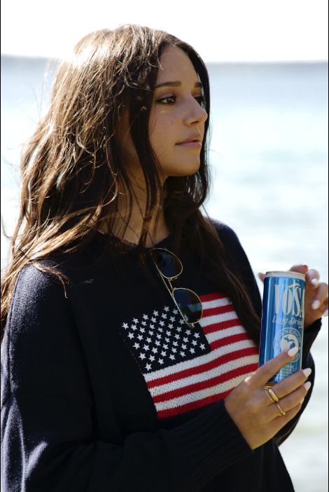 Young woman at the beach holding a can of Voss sparkling water, wearing sunglasses, a shirt with an American flag design, and a dark jacket.