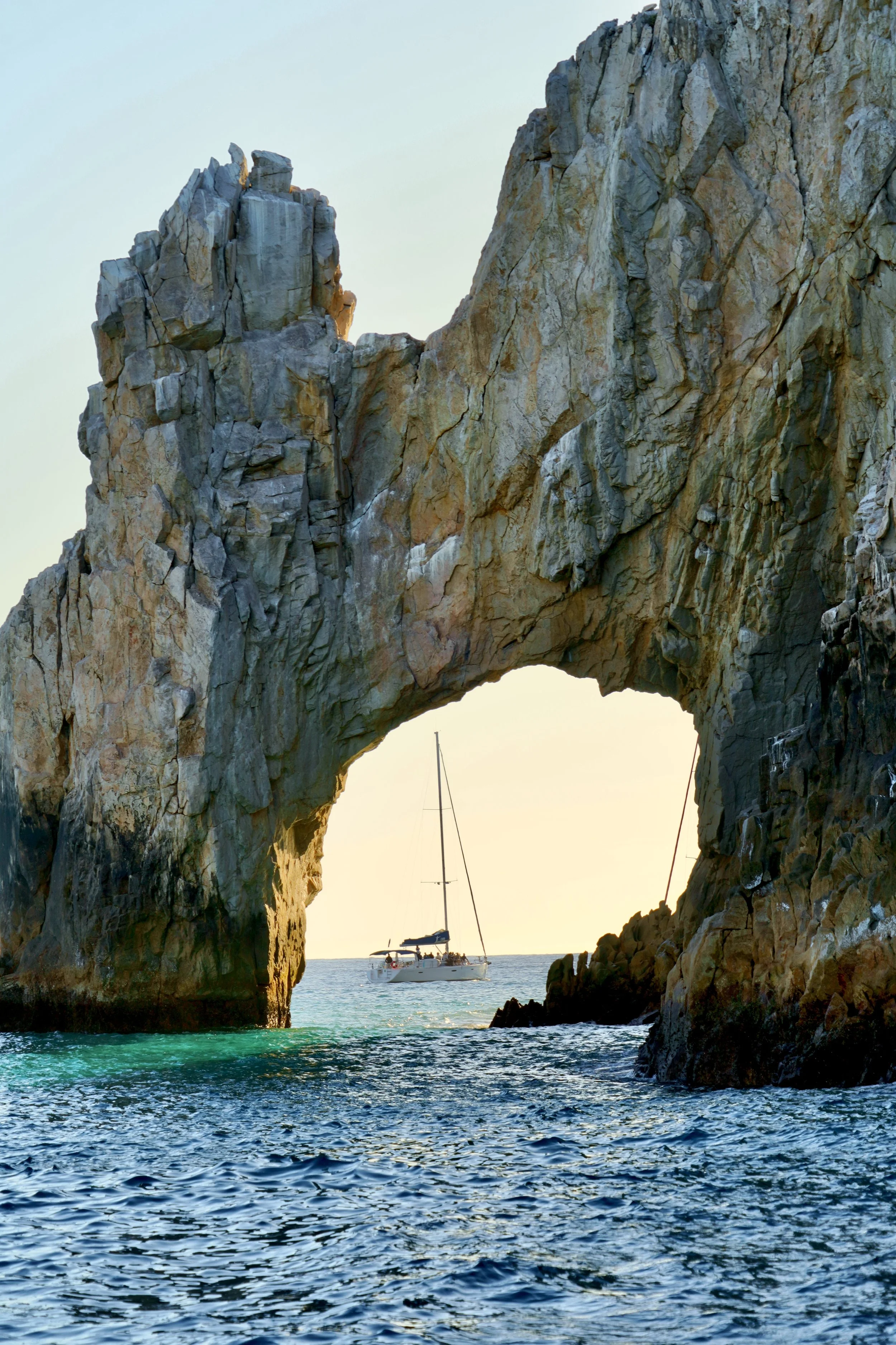 Sailboat passing through a natural stone arch in Cabo San Lucas, Mexico, with ocean and sky in the background.