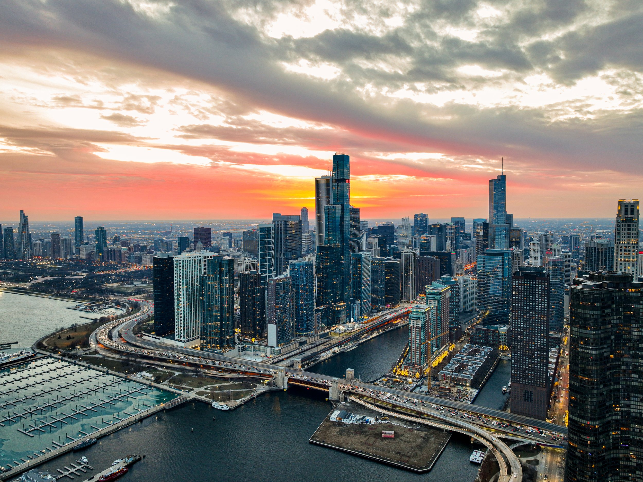 Aerial view of Chicago skyline at sunset with skyscrapers, roads, and Lake Michigan.