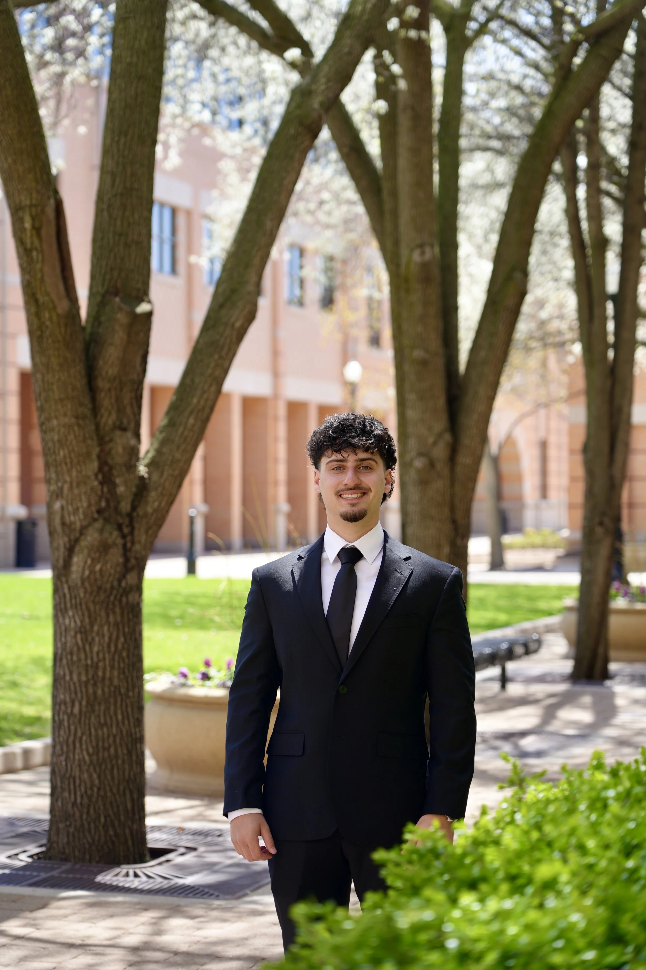 A young man in a black suit and tie standing outdoors in front of trees and a building, smiling.
