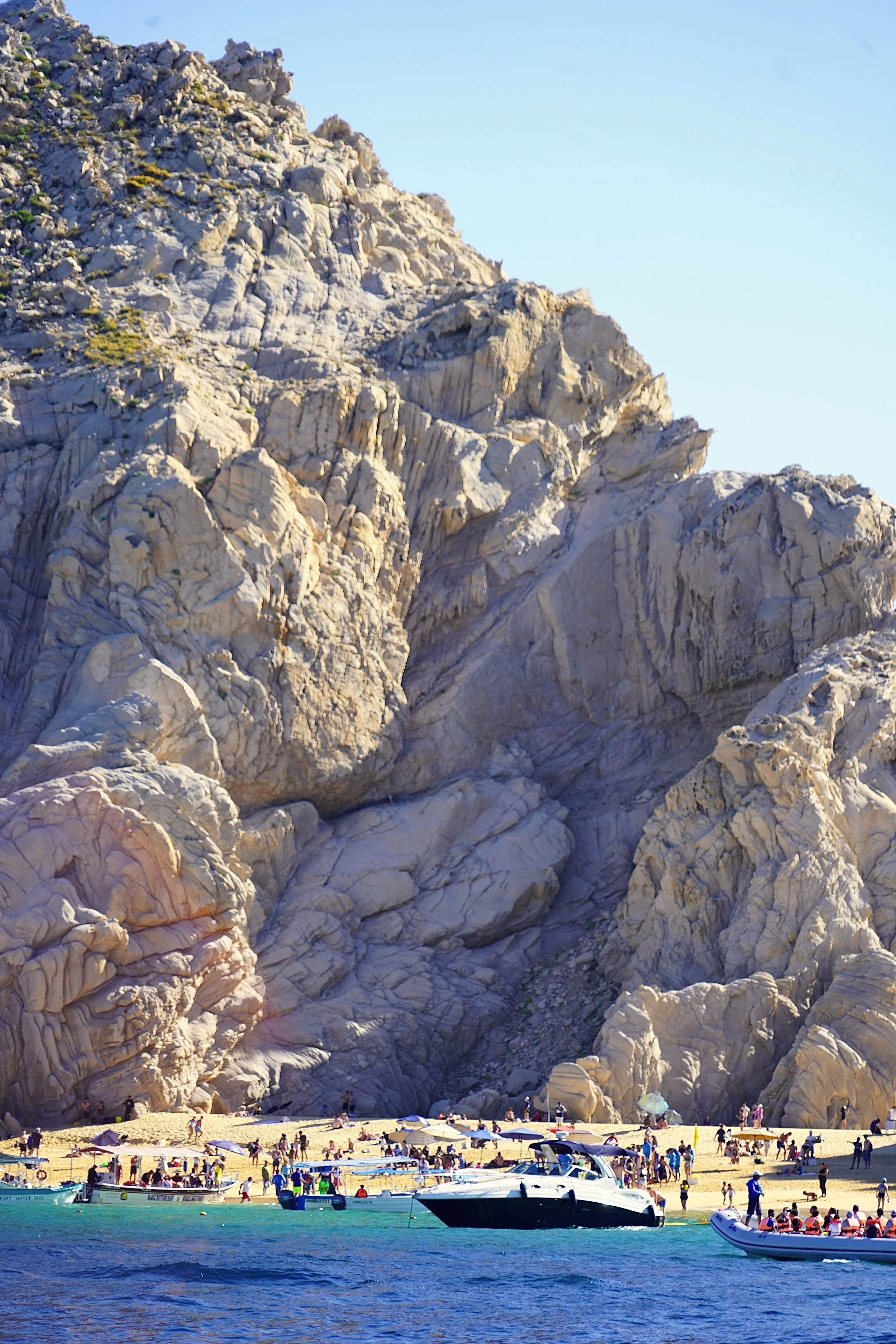 Scenic rocky coastline with a sandy beach, boats, and tourists enjoying the area, under a clear blue sky.