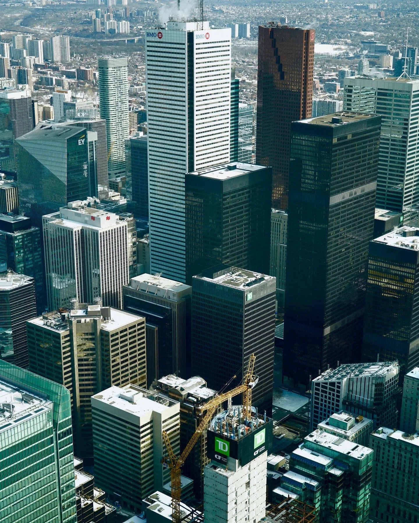 Aerial view of a cityscape featuring modern skyscrapers, including the BMO, EY, and TD Canada Trust towers, with construction cranes visible.