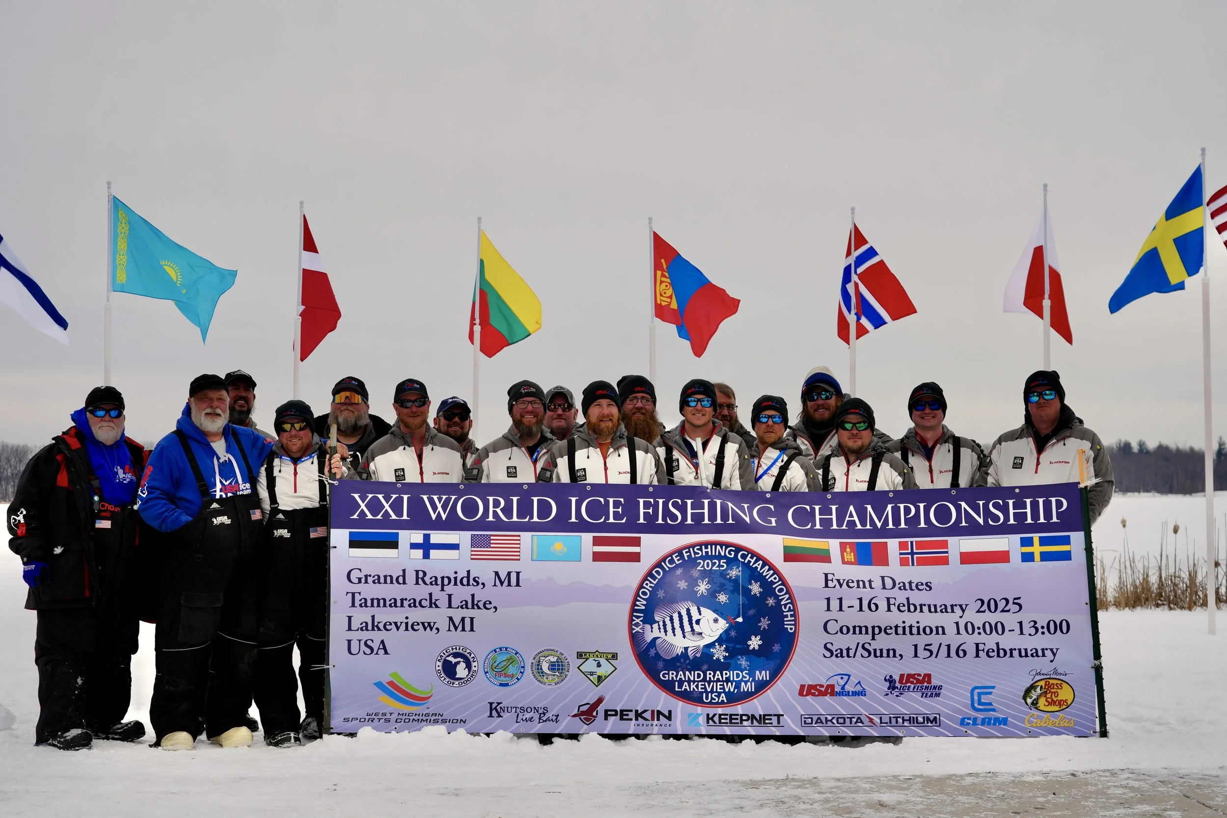 Group of ice fishers holding a banner at the 2025 World Ice Fishing Championship in Grand Rapids, Michigan, with multiple flags flying in the background on a snowy landscape.