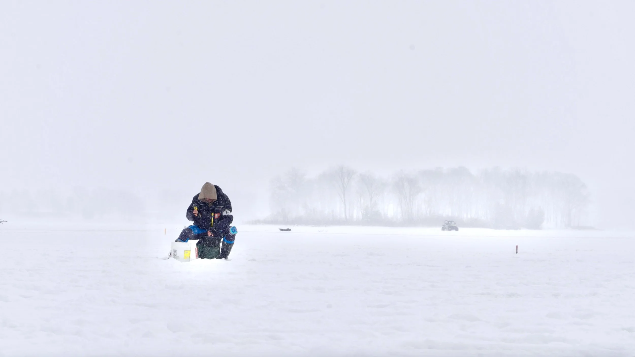 A person ice fishing on a snow-covered frozen lake during a snowy day, with trees and equipment in the background.
