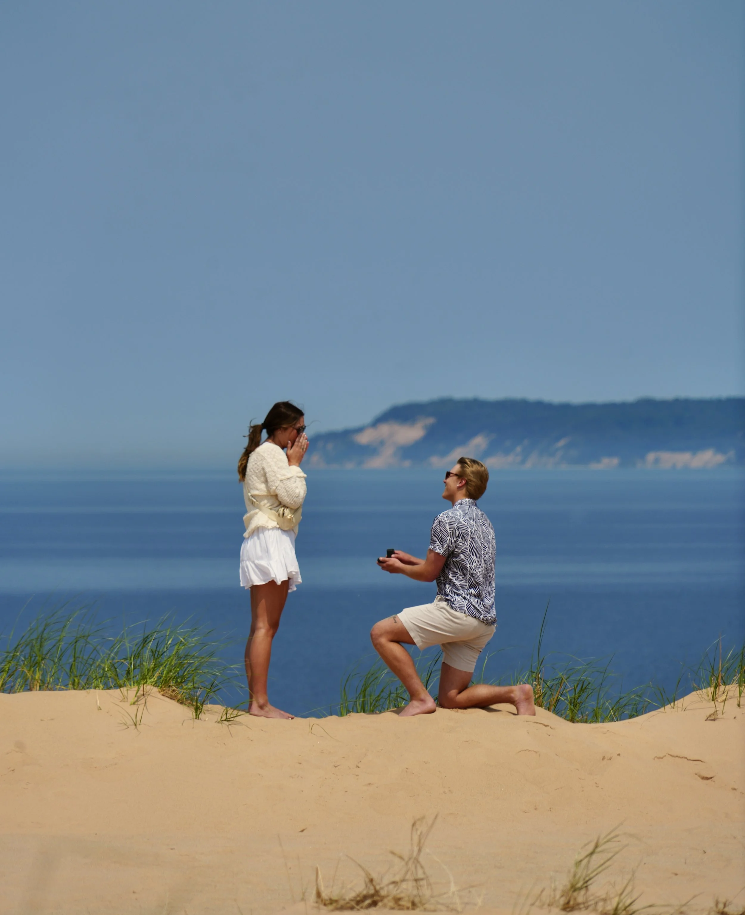 A man proposes marriage to a woman on a sandy beach with grass, blue ocean, and hills in the background, during daytime.