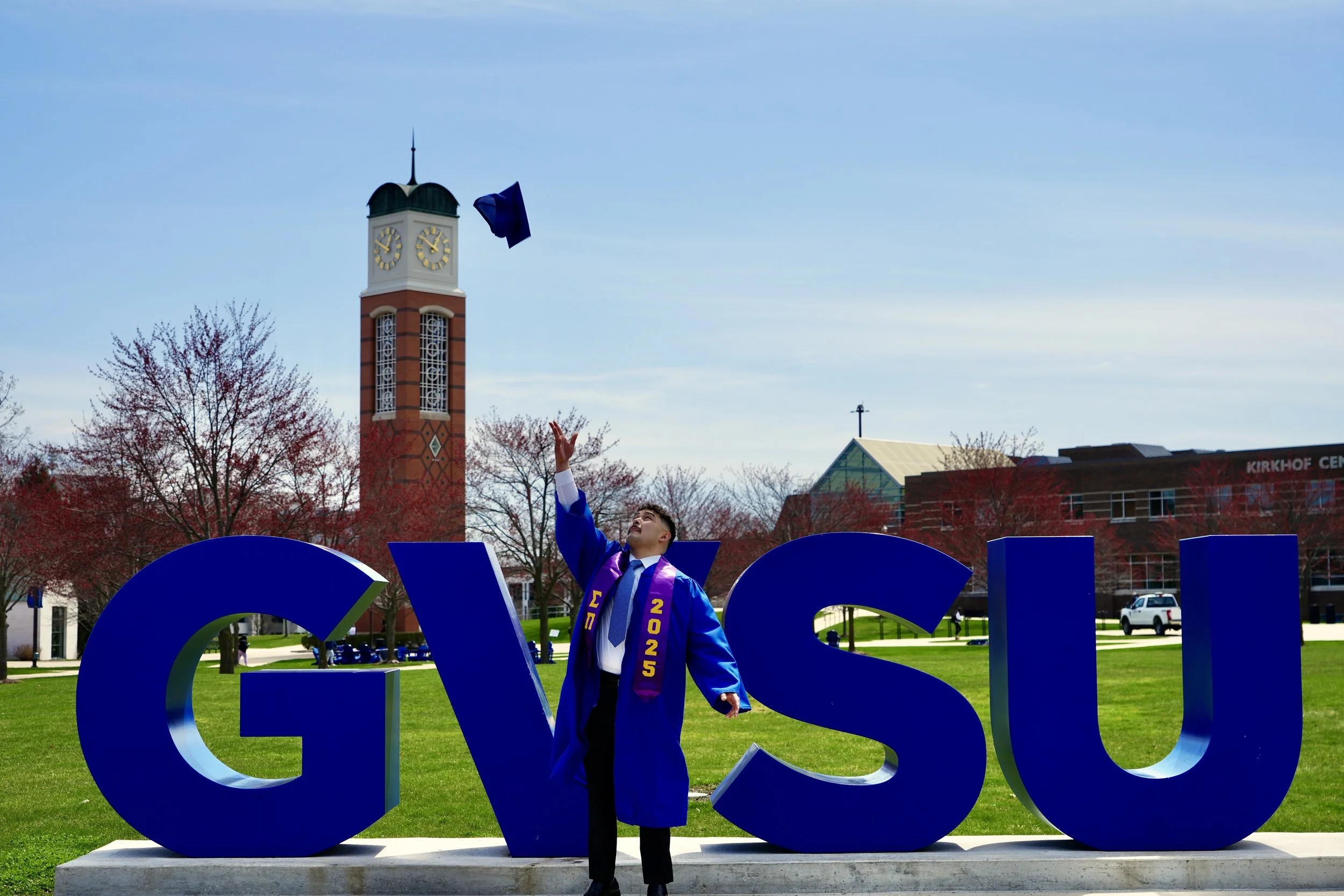 A graduate in a blue cap and gown tossing his cap into the air, standing in front of large blue letters spelling 'GVSU' on a college campus with trees and campus buildings in the background.