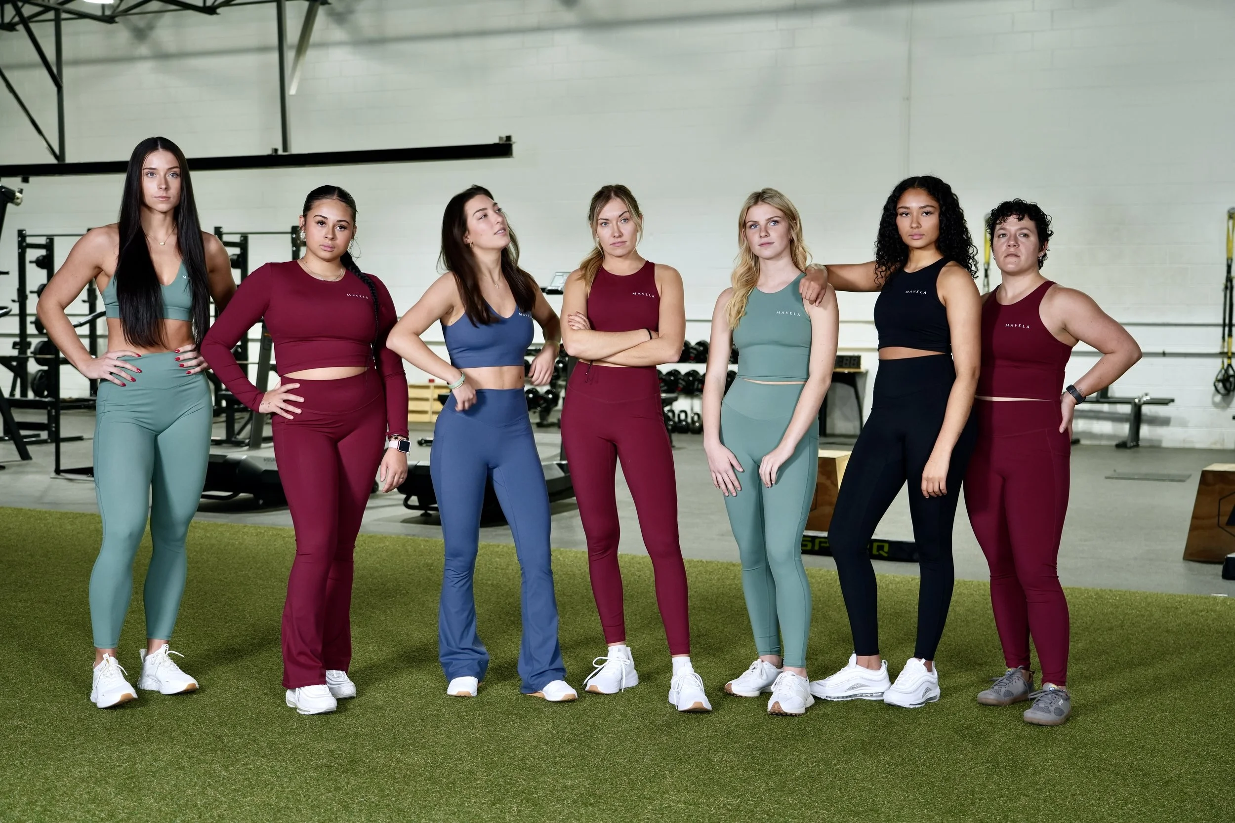 A group of seven women in athletic wear posing together in a gym with workout equipment in the background.