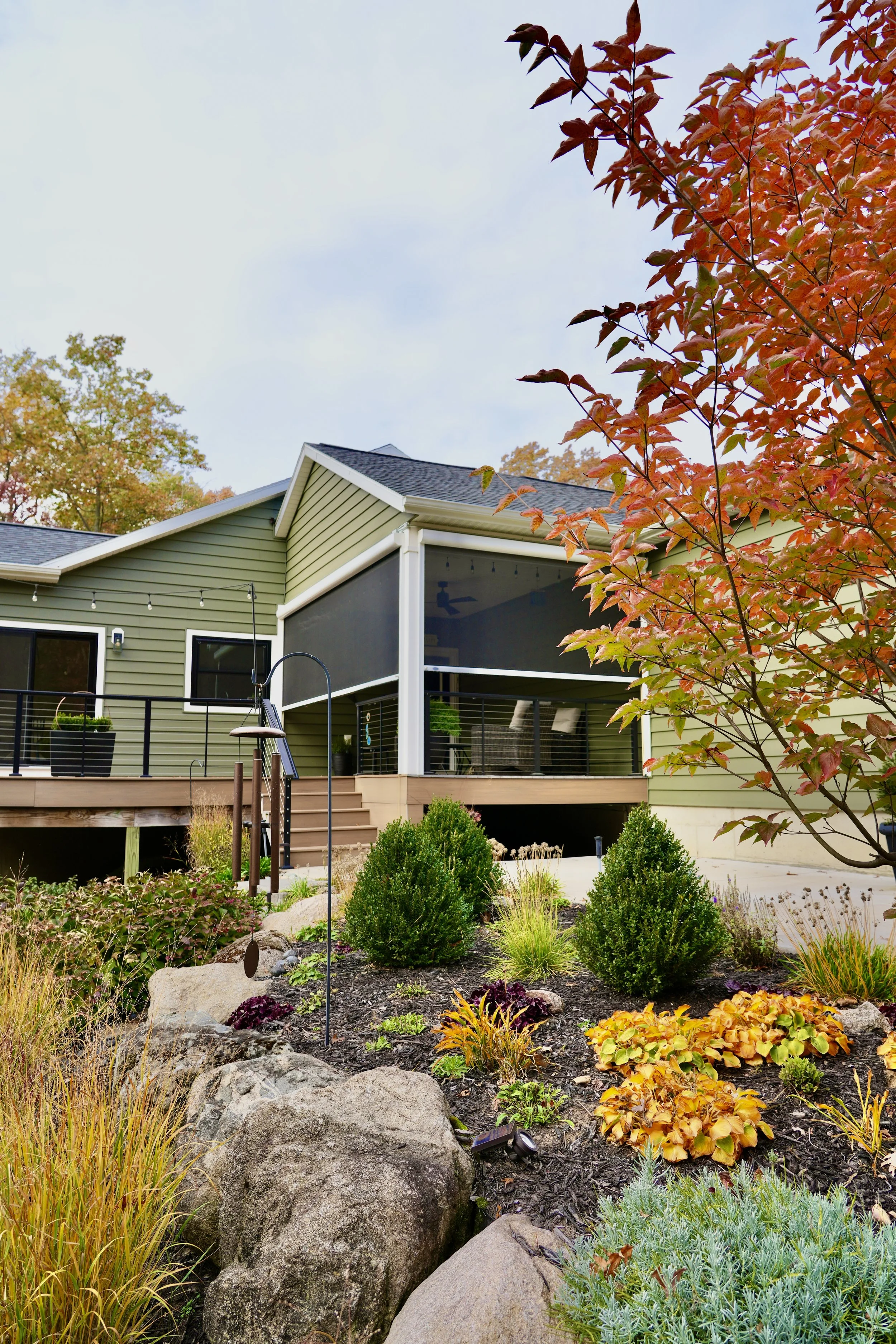 Modern house with green siding and black railings, set against a landscaped garden with rocks, shrubs, and autumn-colored plants. A mature tree with red leaves is in the foreground.