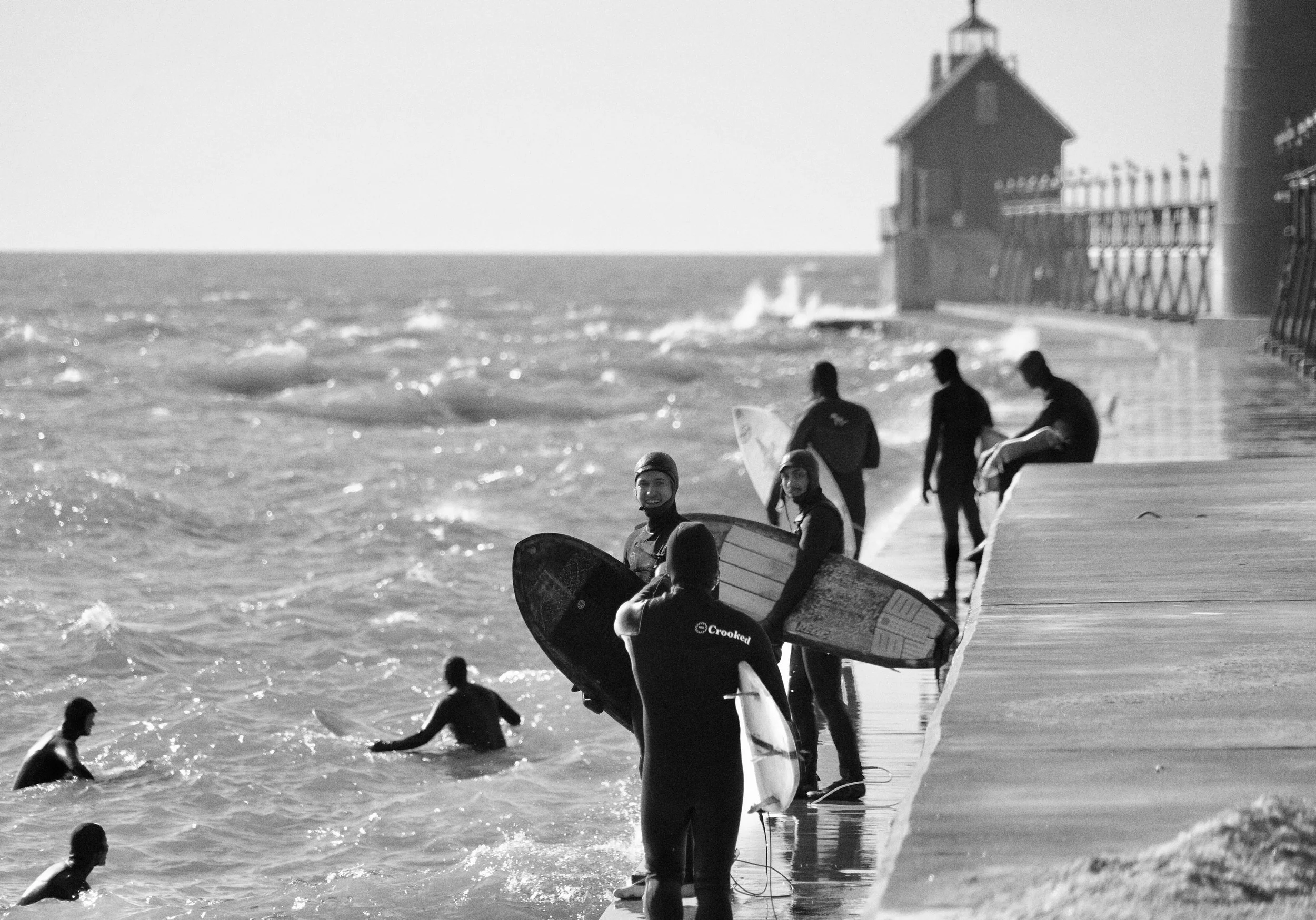 Surfers in wetsuits with surfboards near a pier by the ocean in black and white.