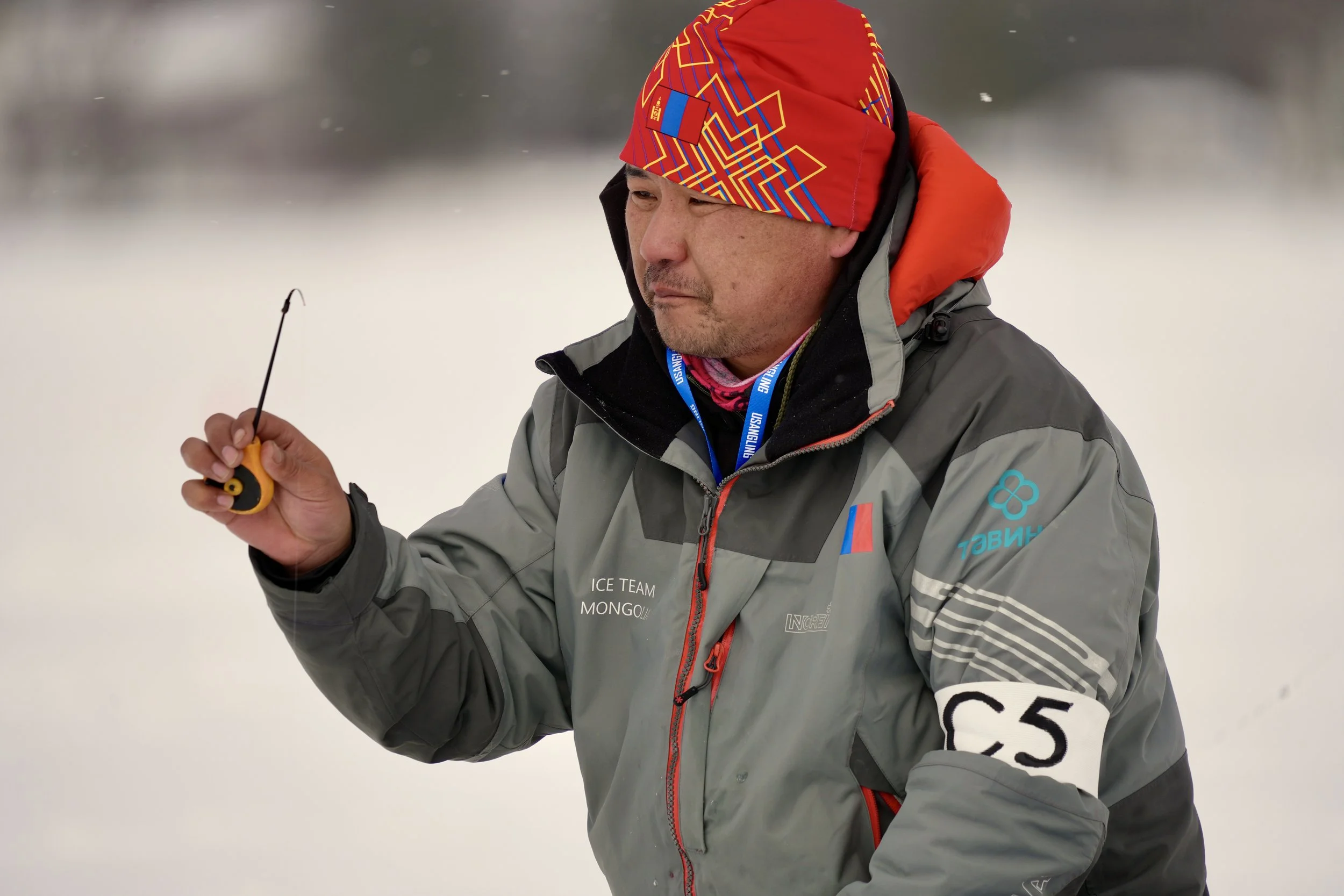 A man dressed in winter gear, including a red patterned beanie, a gray jacket with patches and logos, and a black hood, standing outdoors in a snowy environment, holding a small yellow and black device in his right hand.