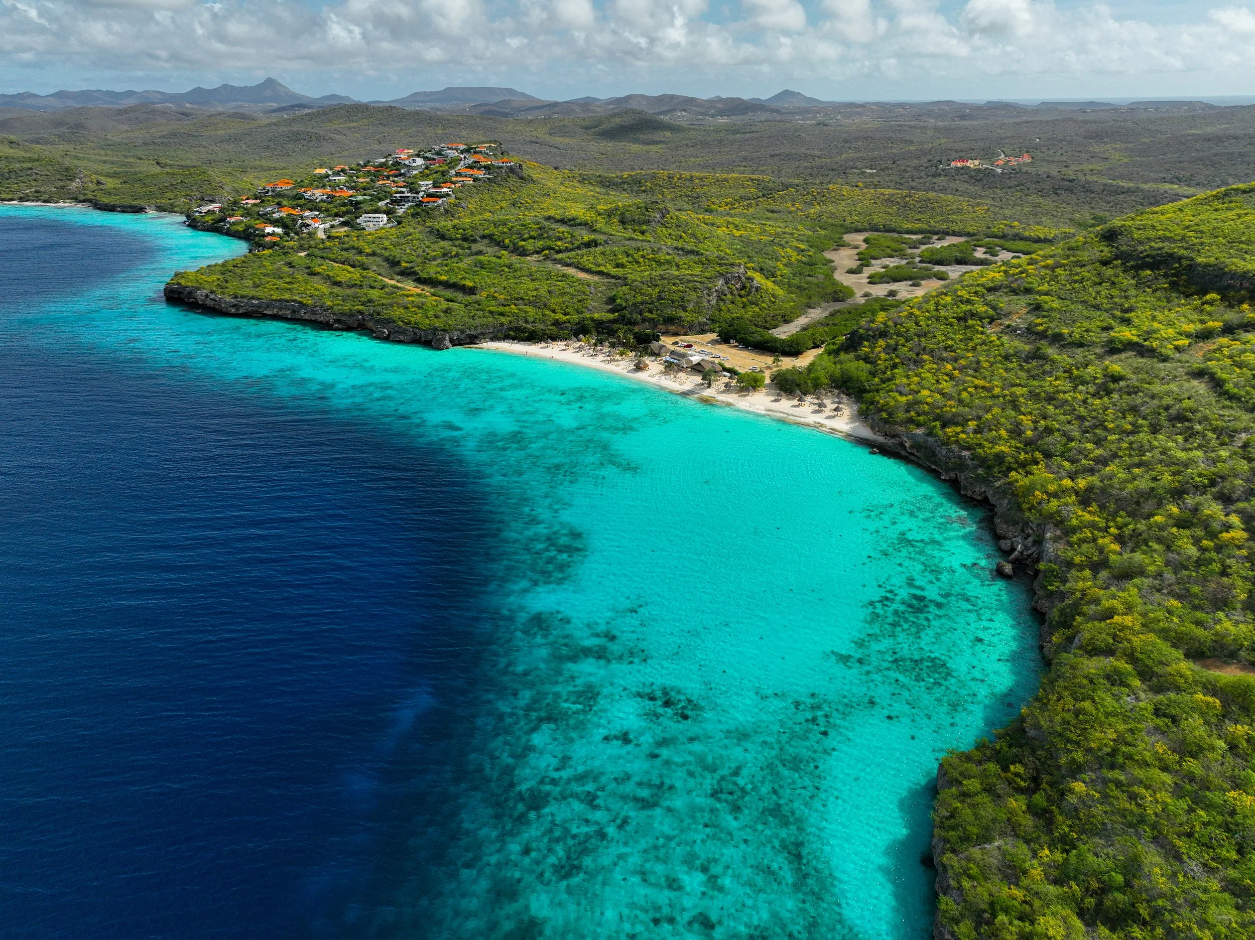 Aerial view of a tropical beach with clear turquoise water, white sandy shoreline, green hills, and a small residential area with orange-roofed houses, under a partly cloudy sky.