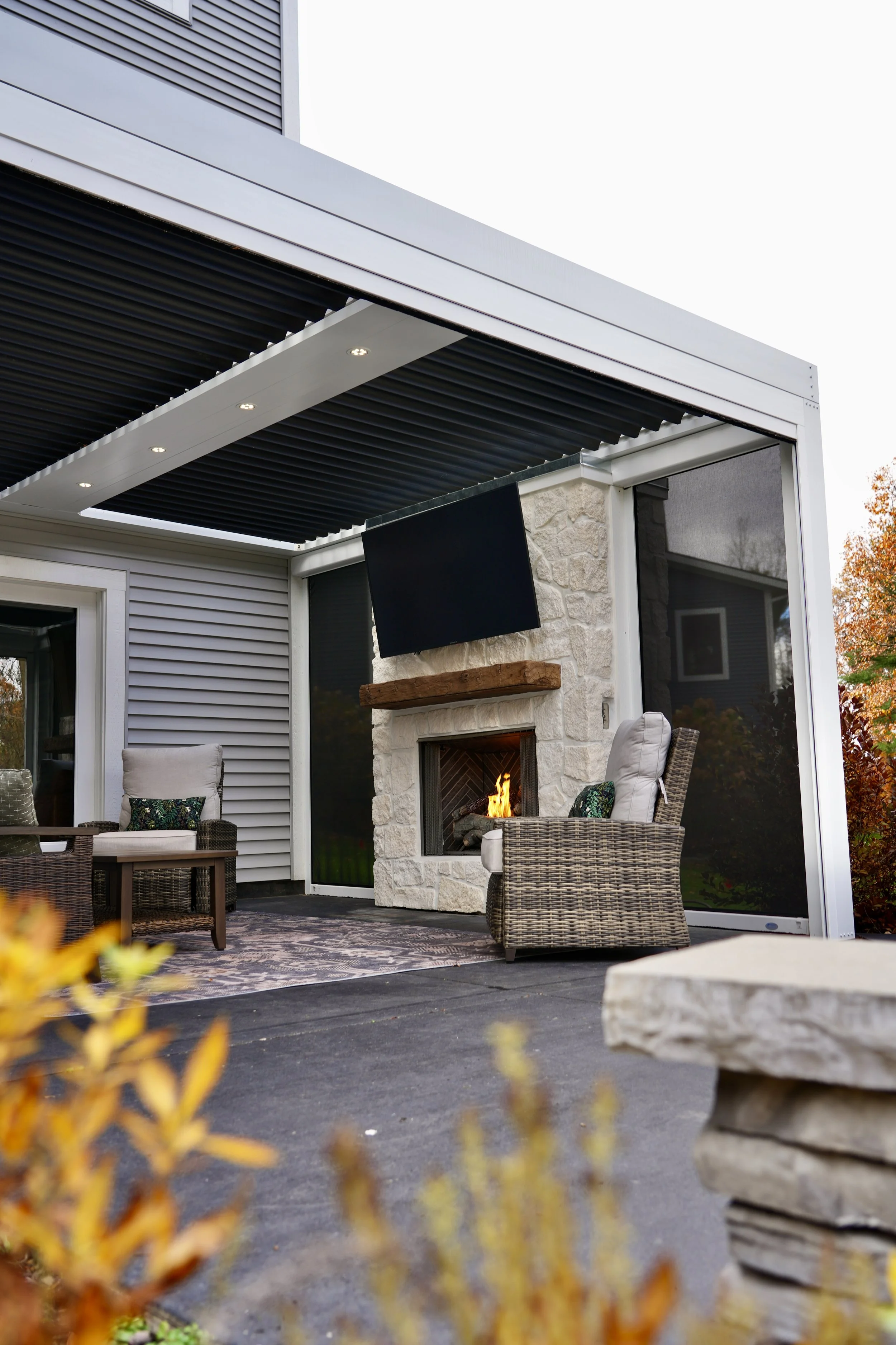 Outdoor patio with a covered seating area, featuring a stone fireplace with a wooden mantel, a mounted TV, and wicker furniture. The area is partially enclosed, with modern siding and large windows.