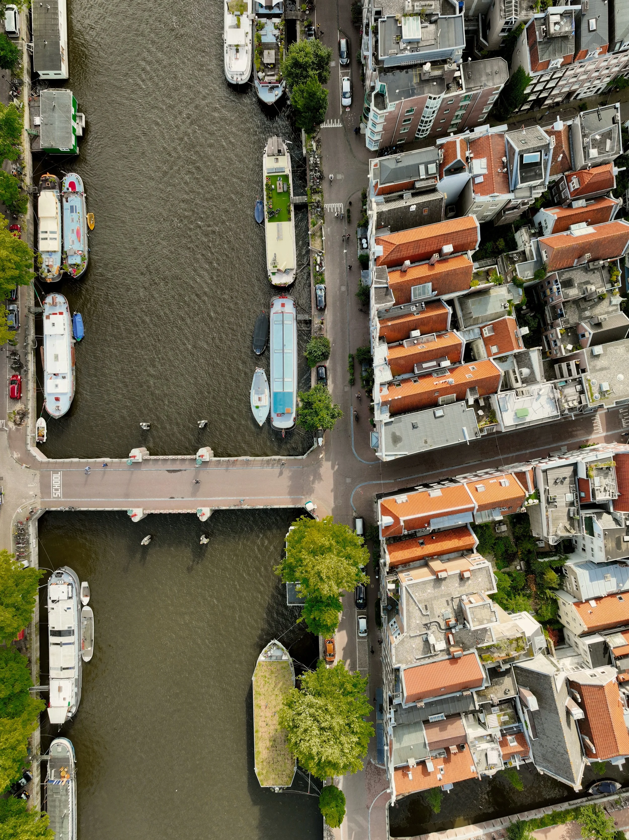 Aerial view of a canal with multiple boats, a pedestrian bridge, and adjacent rows of red-roofed buildings.