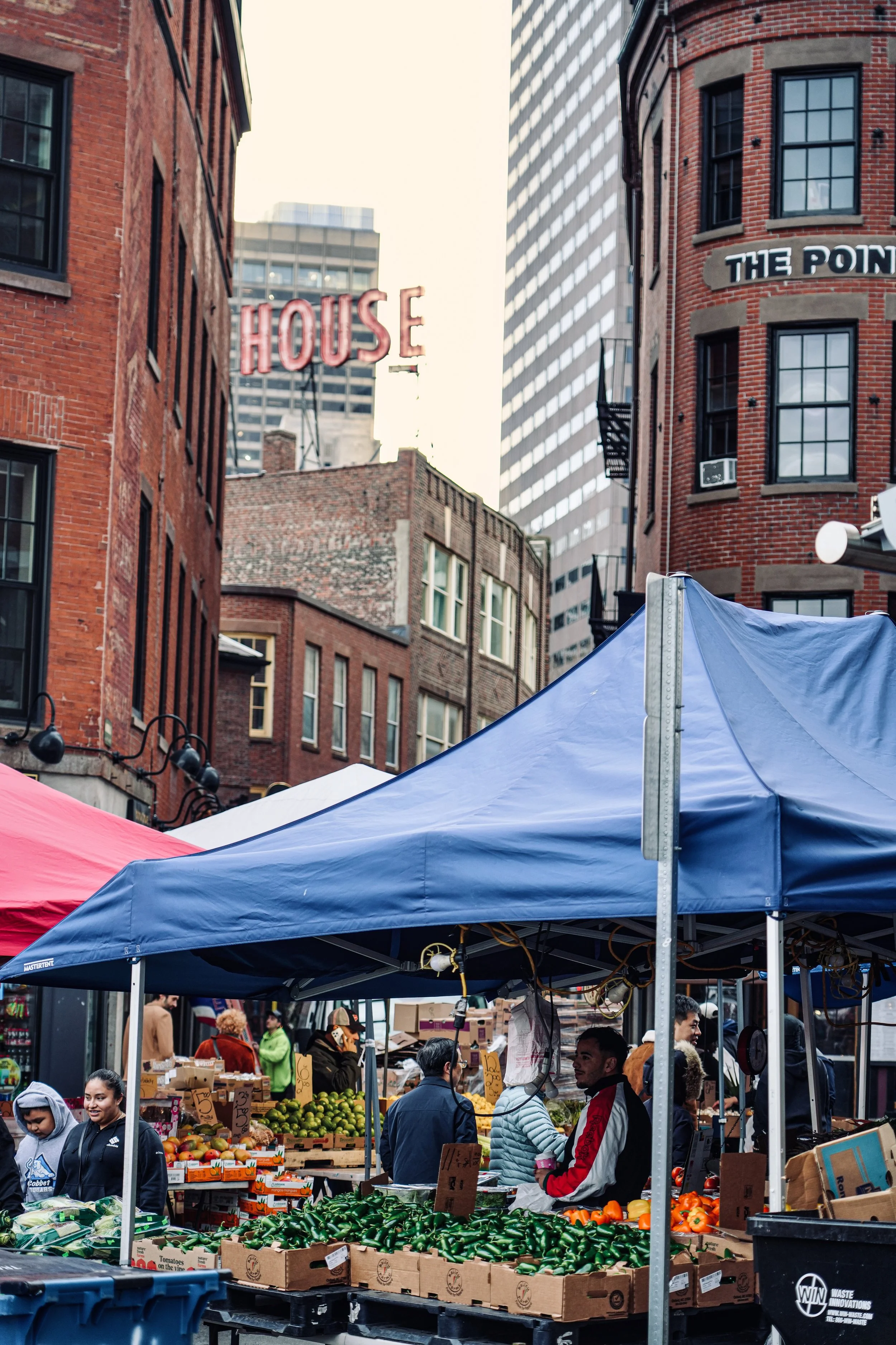 Outdoor market with tents, fresh produce, people shopping, and historic brick buildings in an urban setting.