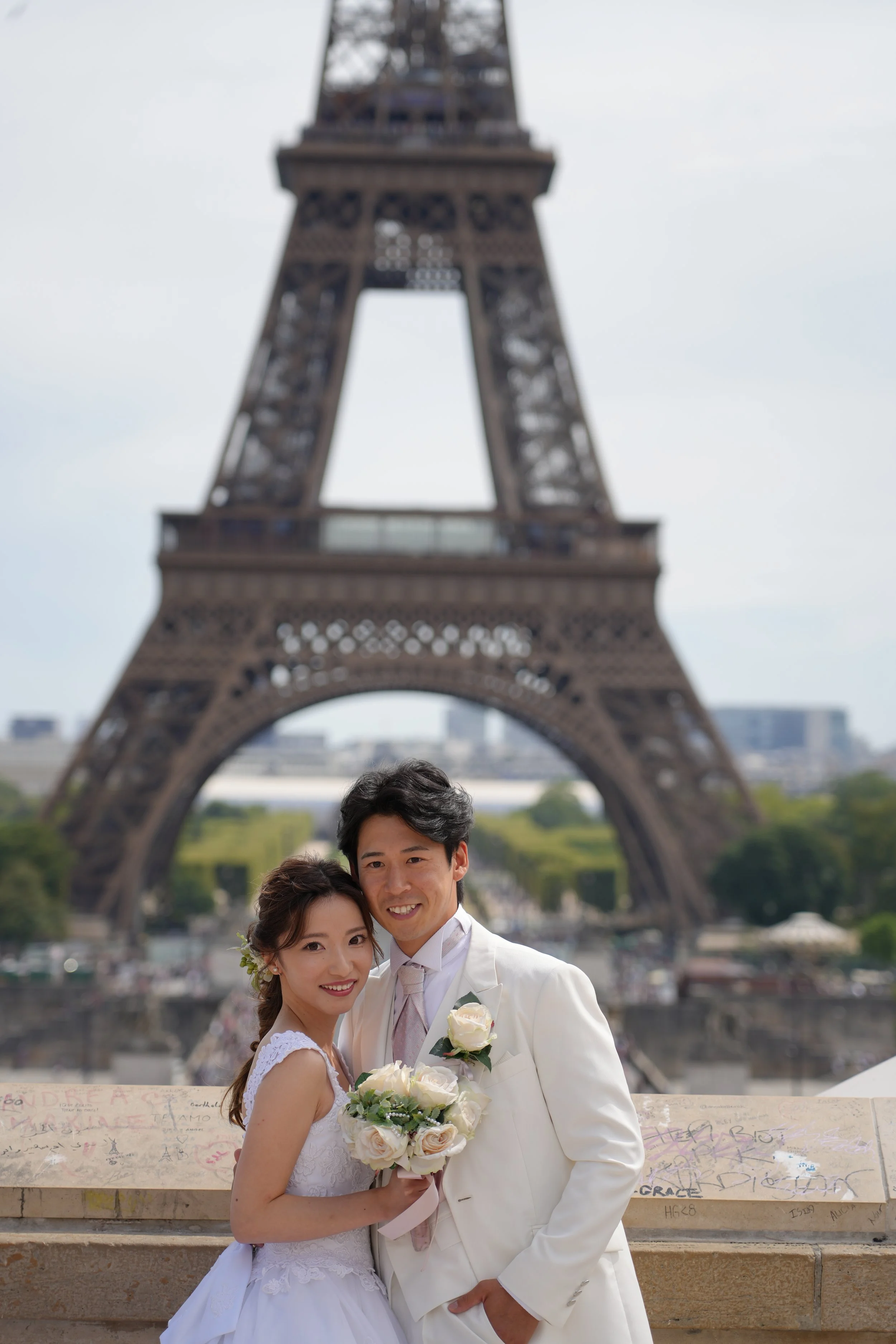 A bride and groom in wedding attire holding a bouquet of white roses, standing in front of the Eiffel Tower in Paris, France.