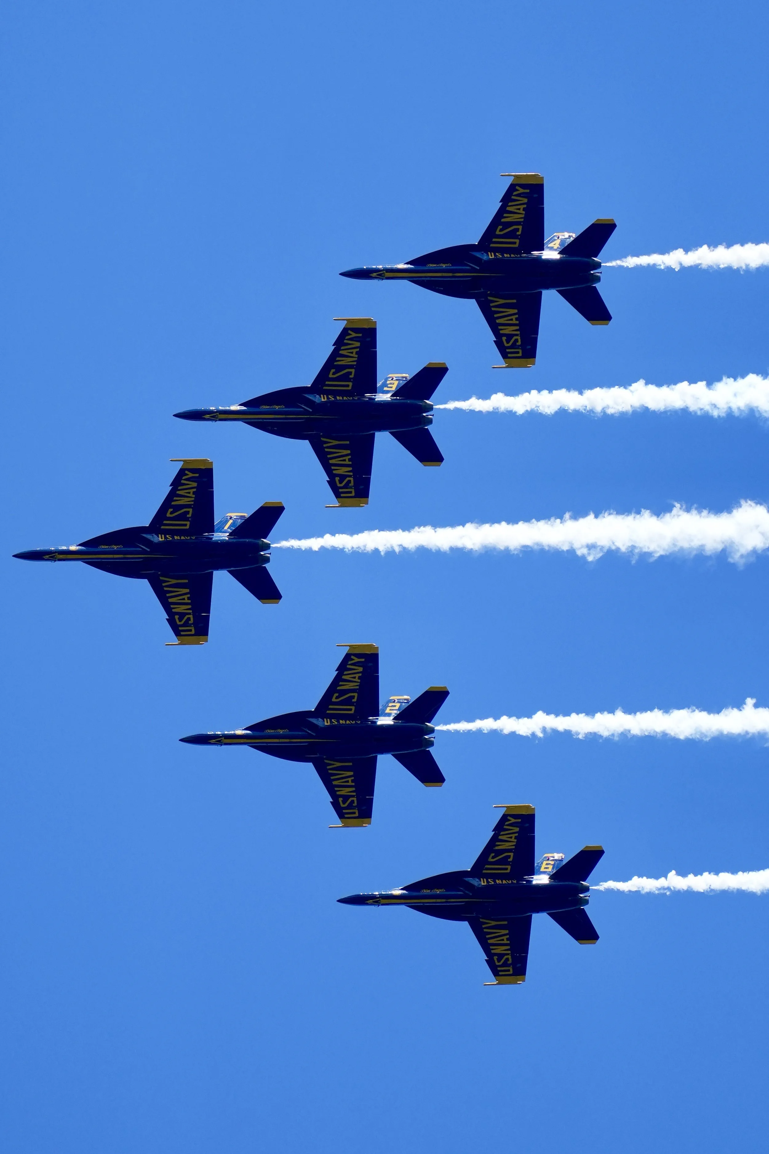 Six U.S. Navy Blue Angels jets flying in formation against a clear blue sky, emitting white smoke trails.