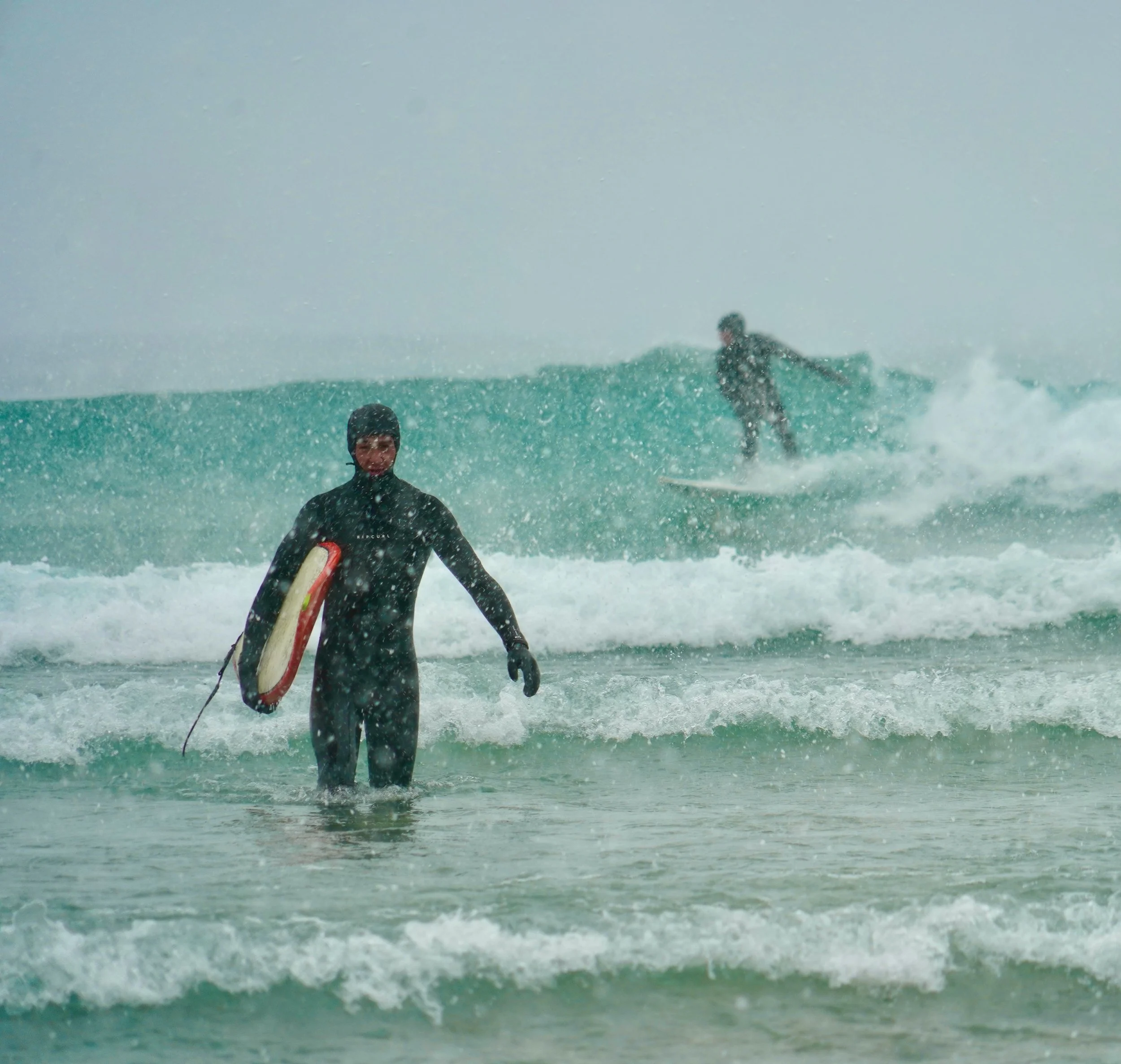 Two surfers in cold weather gear at sea, one holding a surfboard and the other riding a wave with snowfall around.