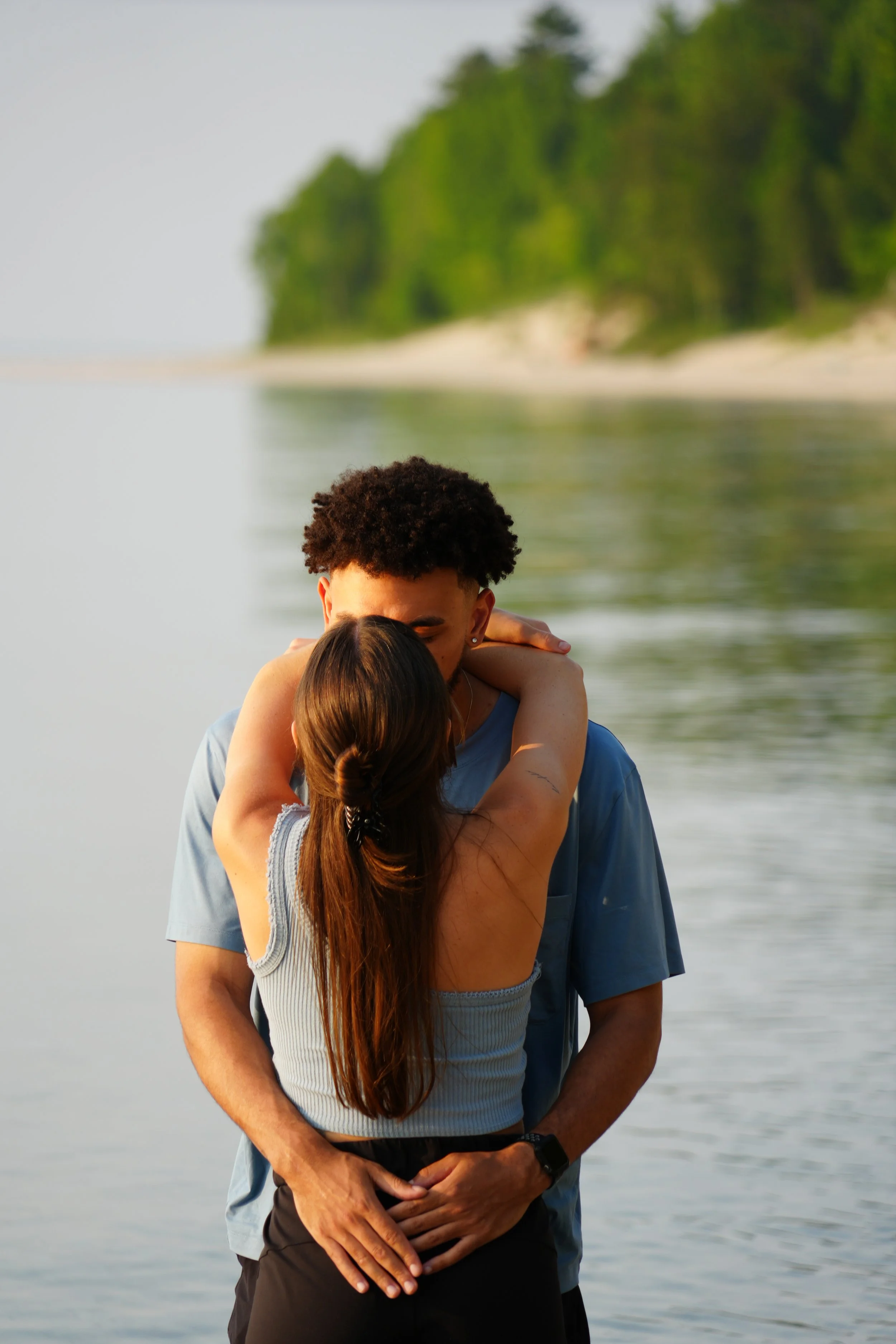 Couple embracing by a lake with trees in the background.