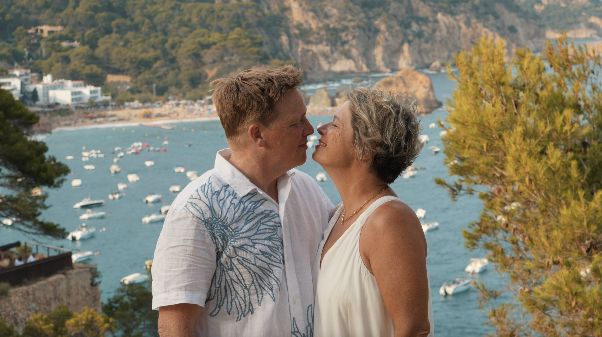 A couple wearing summer attire shares a moment near a picturesque coastal view with boats and cliffs in the background.
