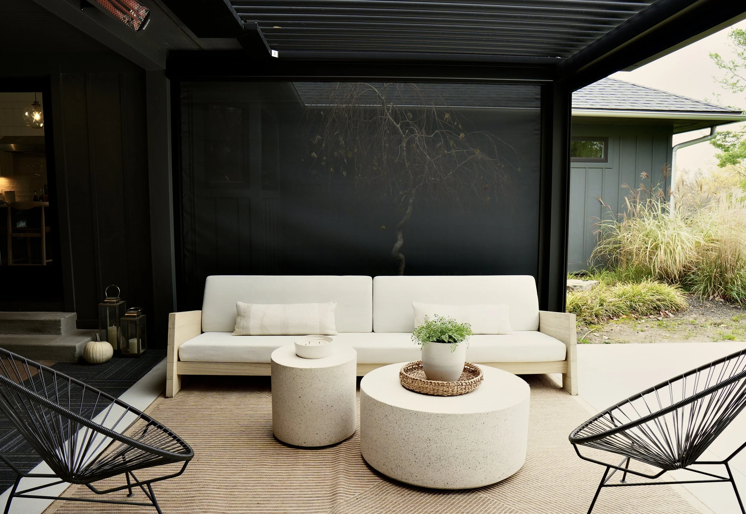 Modern outdoor patio with a white sofa, two round concrete tables, a potted plant, and two black wire chairs on a beige rug. Lanterns and pumpkins in the background.