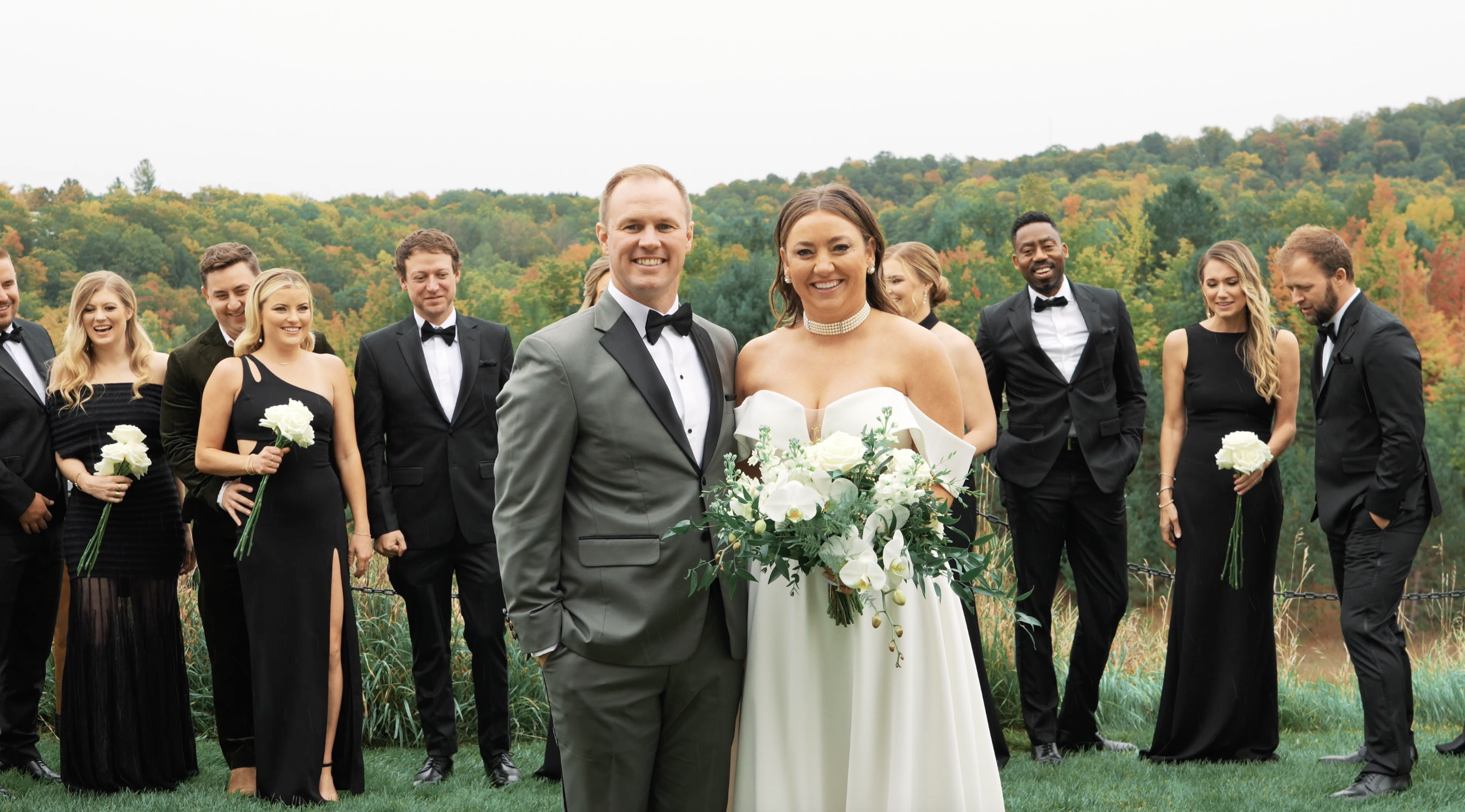 Wedding party posing outdoors, featuring a bride and groom in formal attire surrounded by bridesmaids in black dresses and groomsmen in black tuxedos, with a forested backdrop.