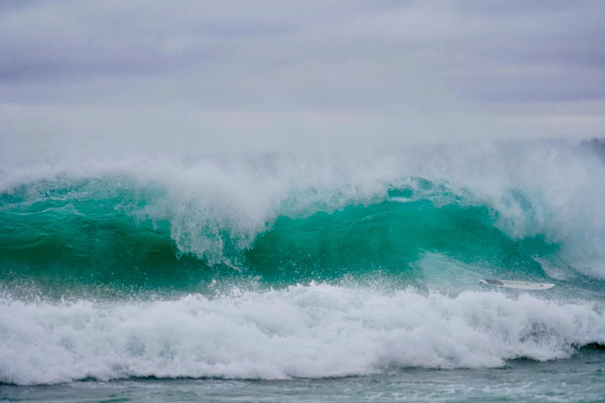 Large ocean wave crashing with surfboard in foreground on a cloudy day.