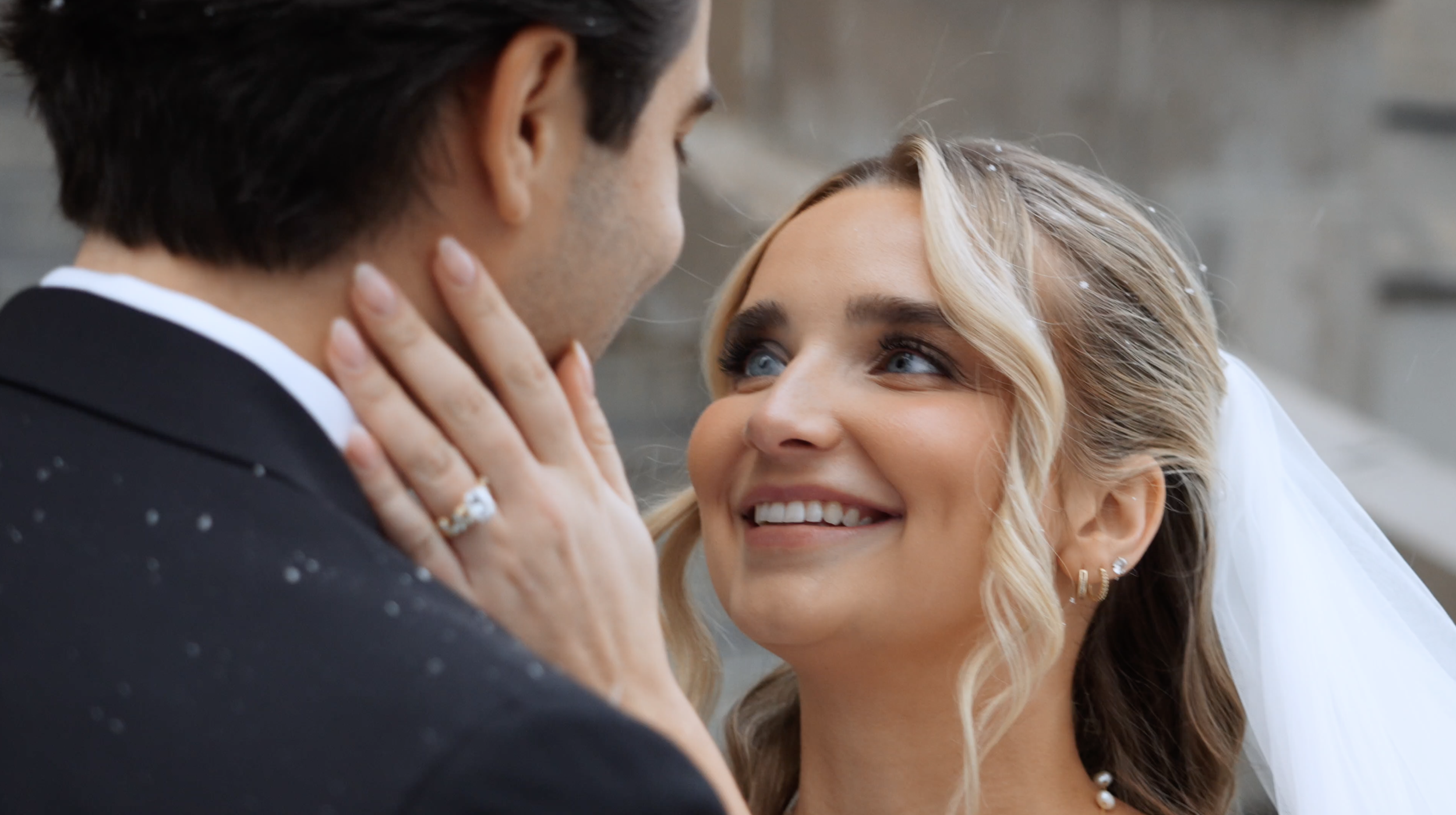 A bride and groom share an intimate moment on their wedding day, gazing into each other's eyes; the bride has blonde hair, blue eyes, and a veil, while the groom has dark hair and is wearing a suit.