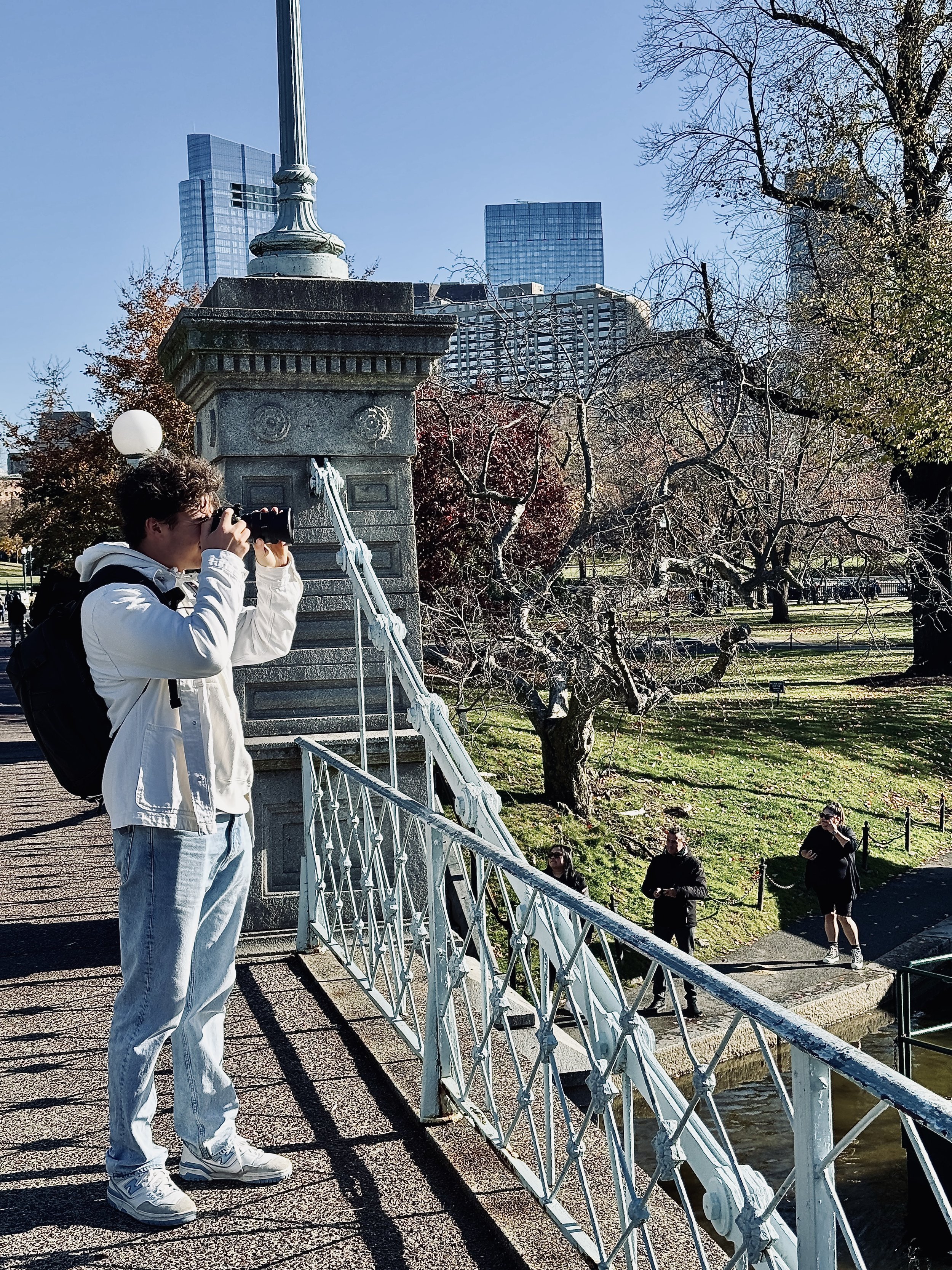 Person taking photos on a bridge in an urban park with city buildings in the background.