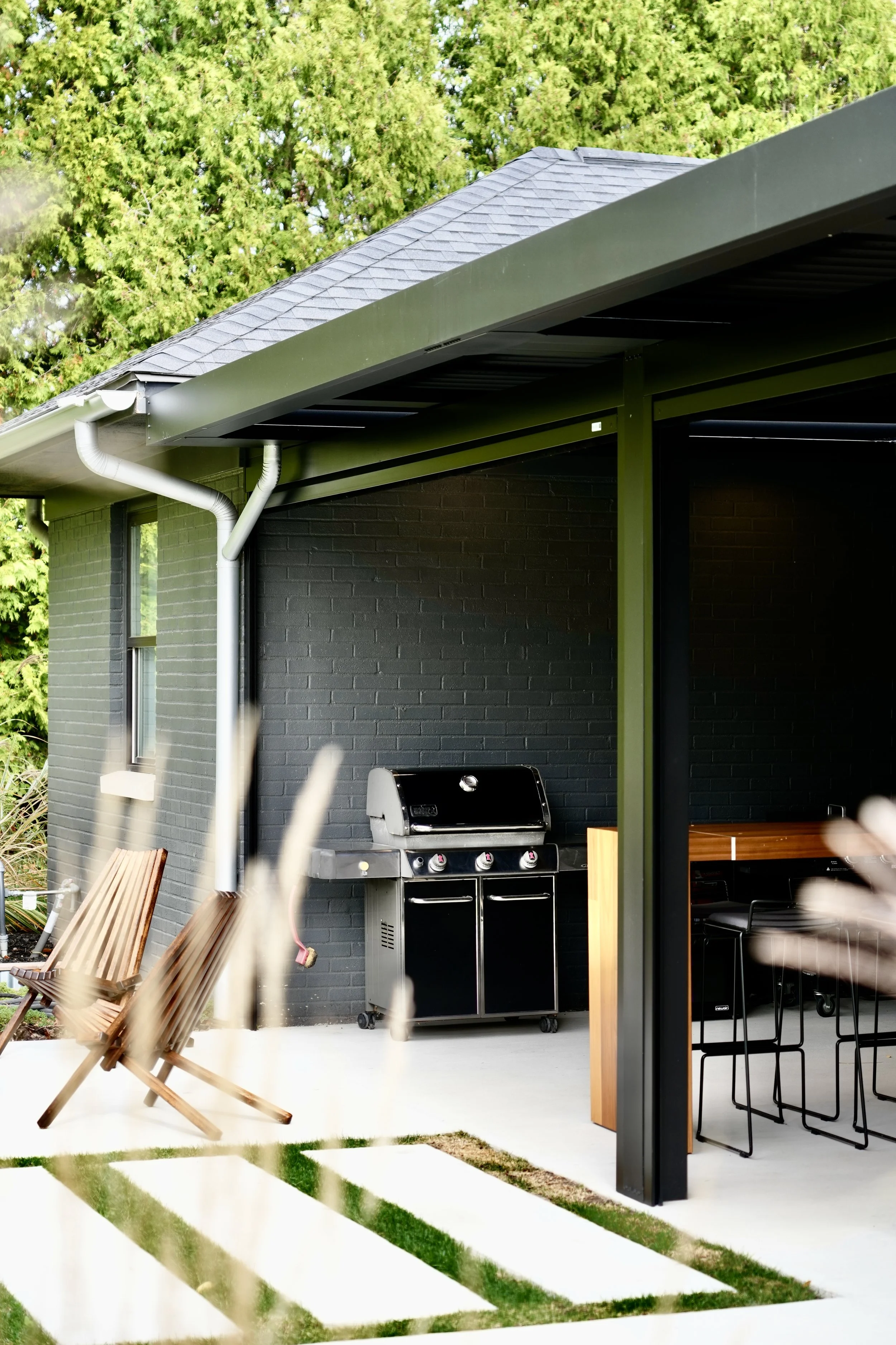 Modern outdoor patio with black grill, wooden chairs, and bar-style table set, surrounded by greenery.