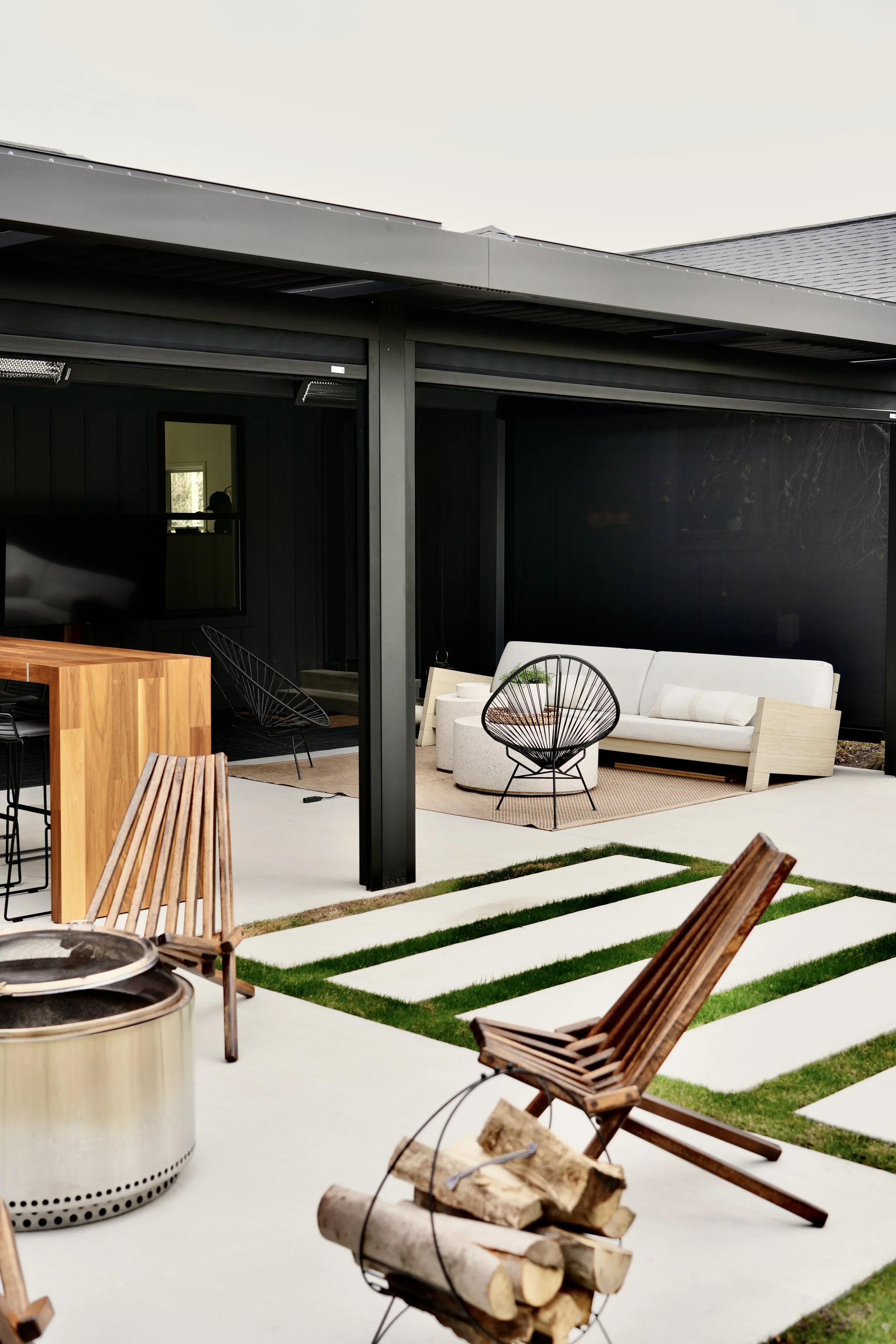 Modern outdoor patio with wooden chairs, a metal fire pit, and stacked firewood. Features a wooden dining table, black wire chairs, and a white sofa under a covered area. Grass strips with concrete pathways.
