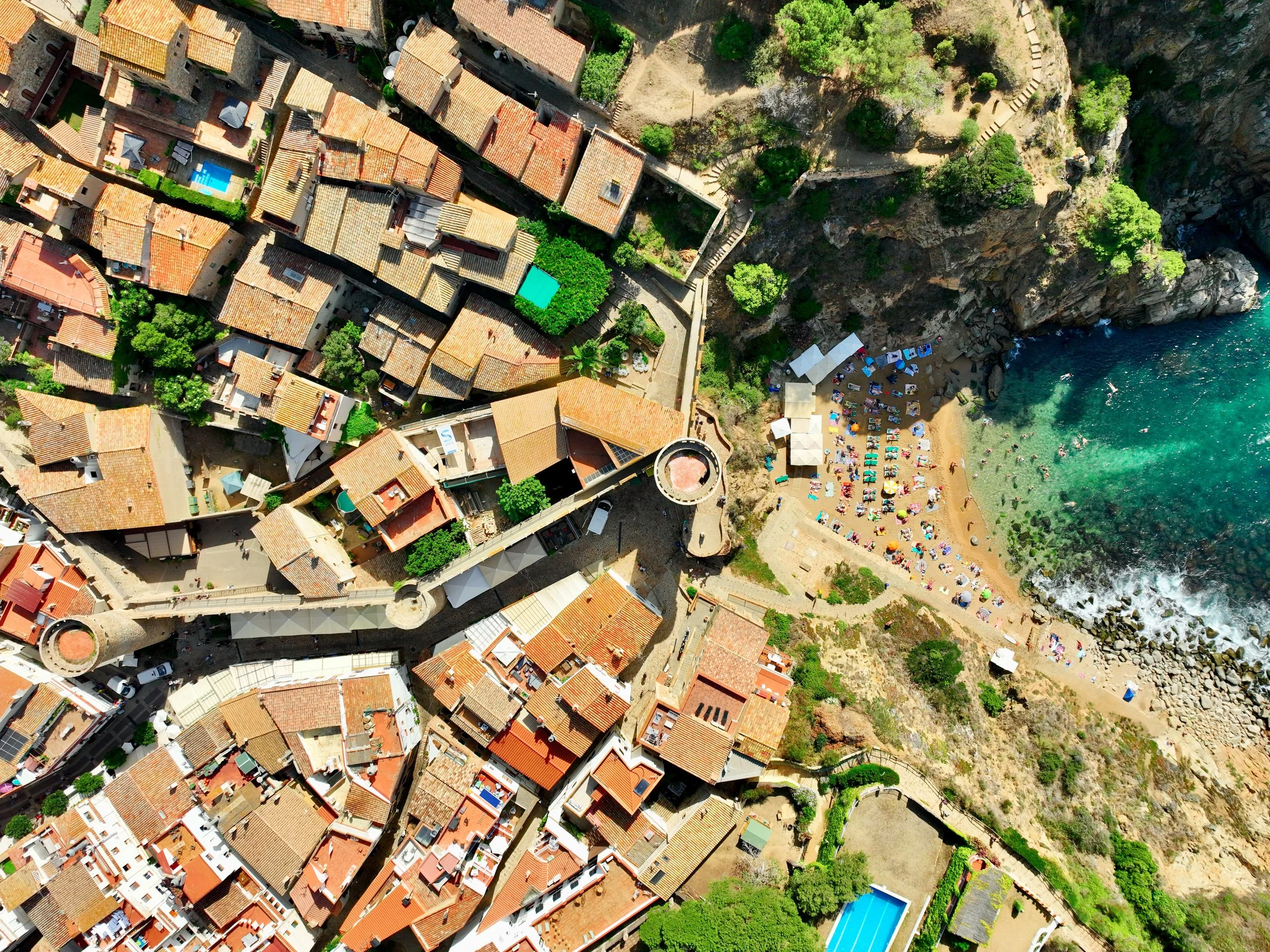 Aerial view of a coastal town with red-tiled rooftops, medieval walls, and a beach. Many people are sunbathing and swimming. The sea is clear and turquoise.