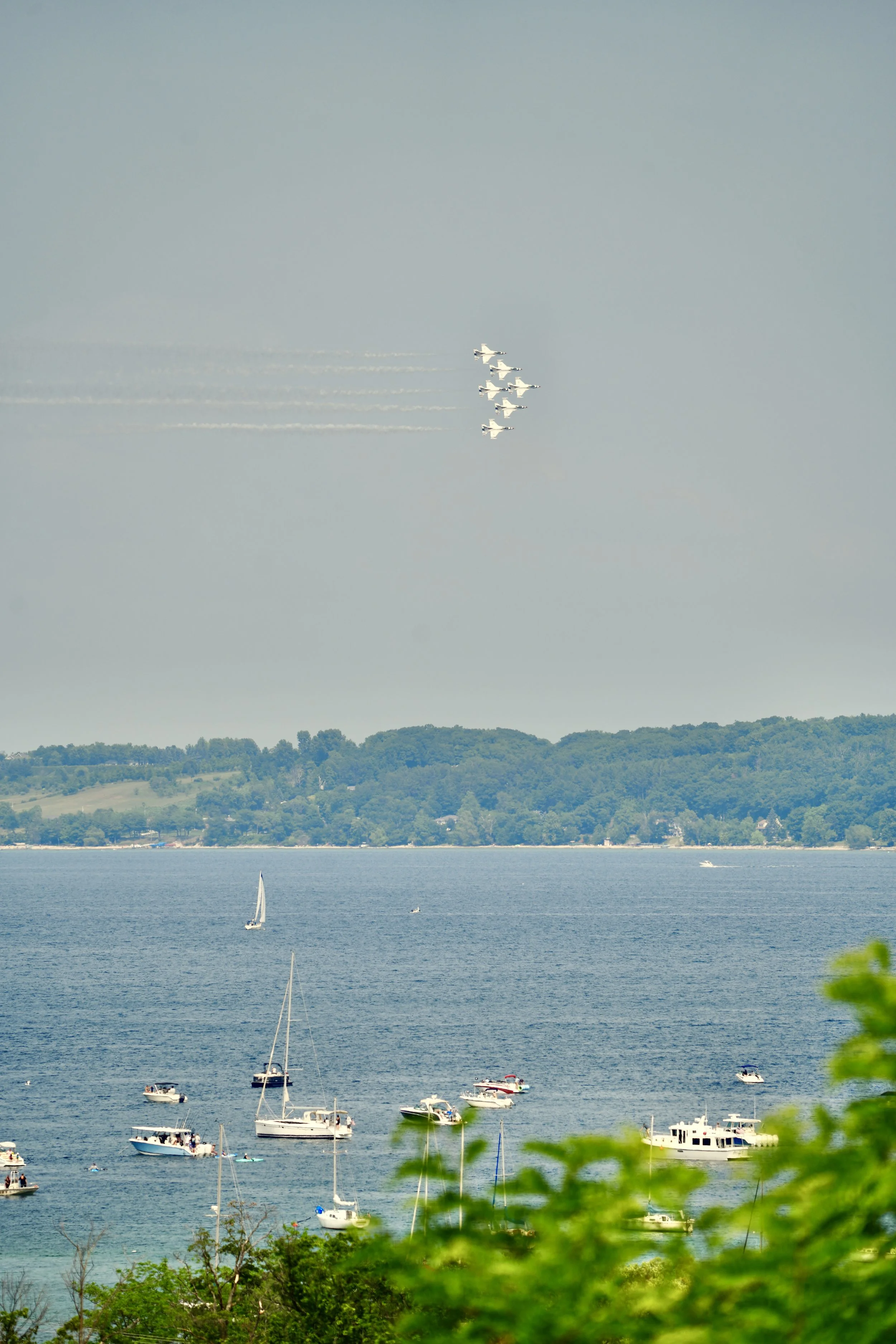 Aerial view shows a formation of six fighter jets flying in the sky, leaving smoke trails. Below, a body of water with multiple sailboats and motorboats is visible. Trees and a distant forested shoreline are in the background.